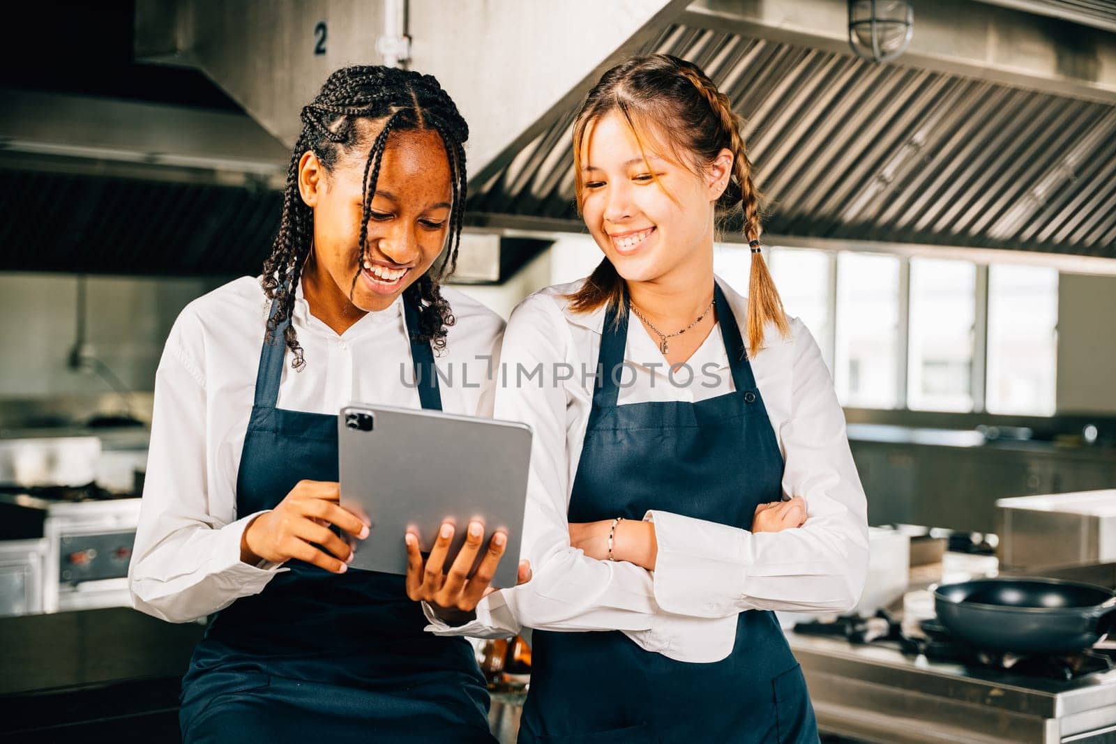 Two famous chefs in stainless steel kitchen discuss video blog using tablet. Professionals smiling standing reading searching. Modern technology in restaurant industry holding device. by Sorapop