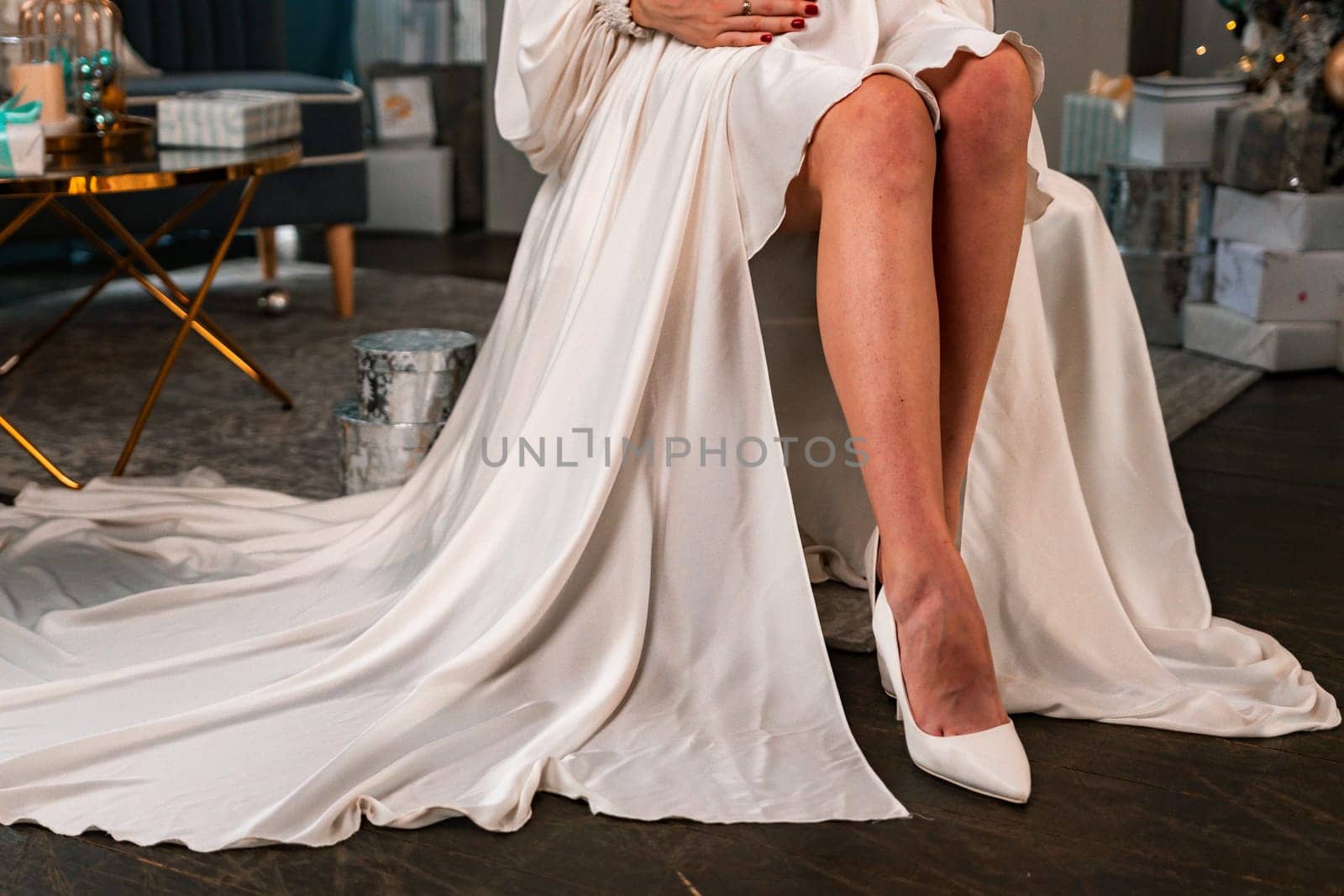 Pregnant Women legs christmas tree. Cropped photo of beautiful elegant female legs in white high-heeled shoes and a white dress, a woman sits against the background of a Christmas tree with gifts. by Matiunina