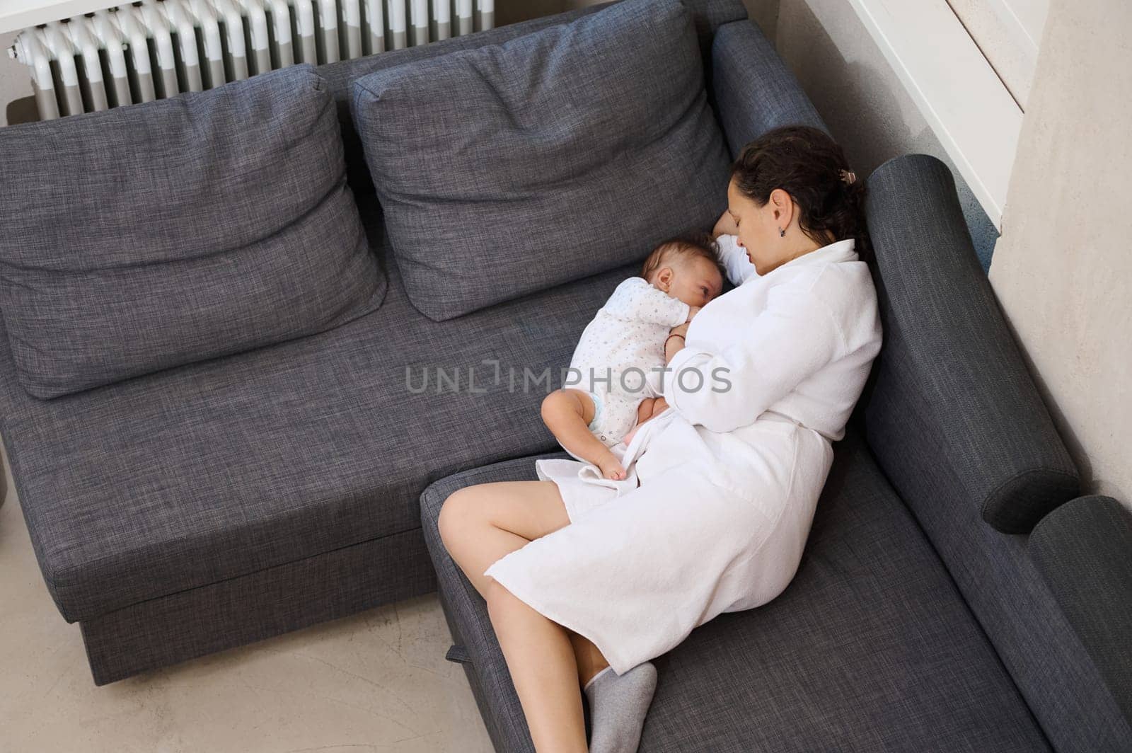 View from above of a mother breastfeeding her baby, lying together together on the sofa. Young woman in white waffle bathrobe, relaxing on the couch, near her newborn baby suckling the mother's milk