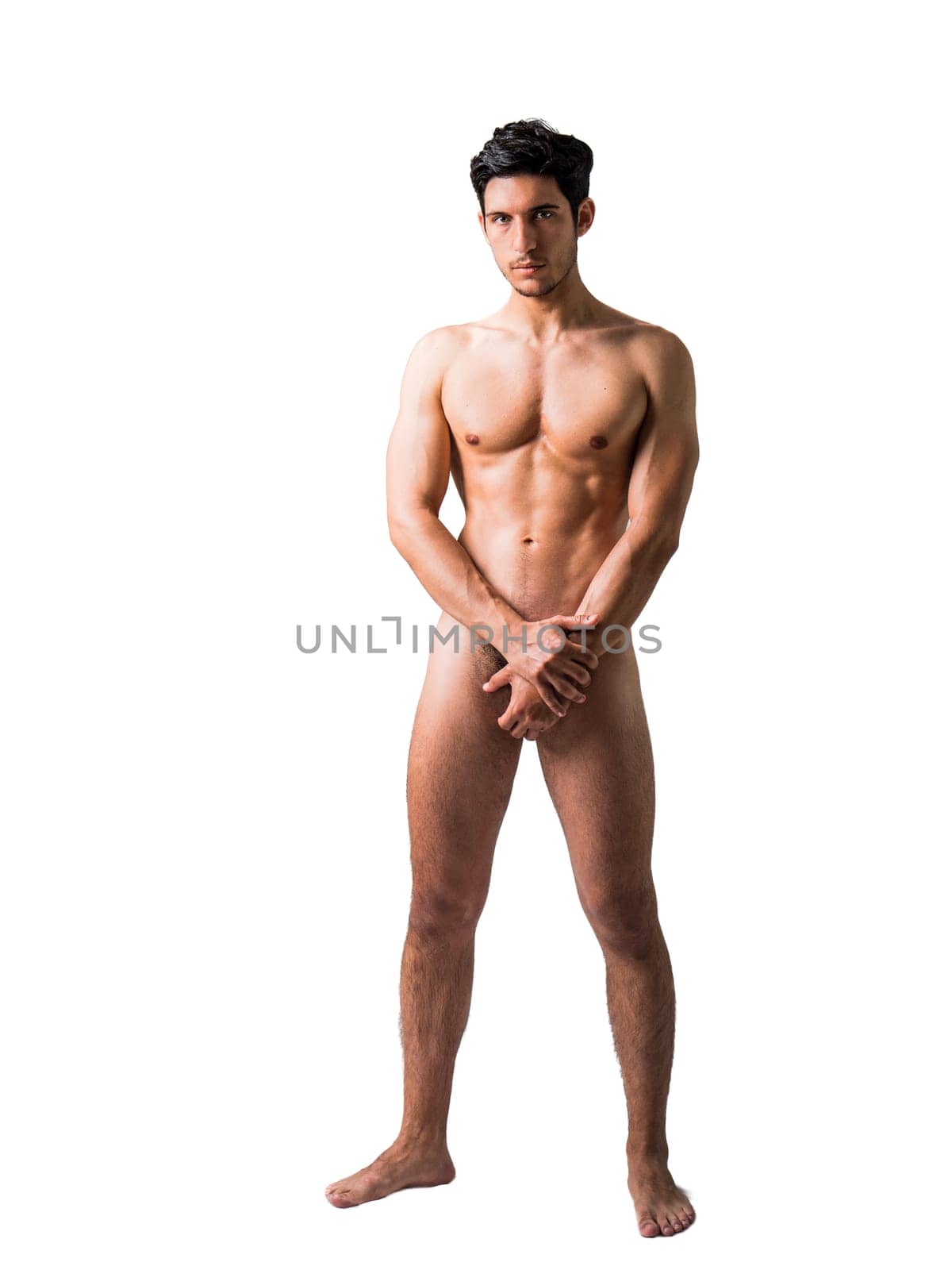 Totally naked athletic handsome young man, looking straight in a muscular tension pose, isolated on white background in studio. Full length shot of naked handsome young man with languishing look covering crotch with a hands, light neutral background