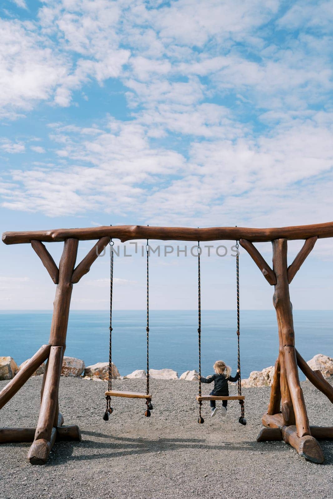 Little girl sits on a large wooden swing on a seashore and looks to the side. Back view. High quality photo