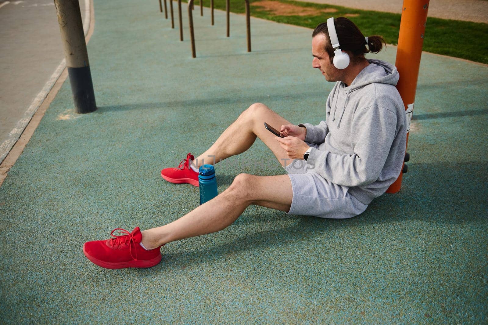 Athletic man in headphones, checking mobile app on his smartphone while resting after workout the urban sports ground by artgf