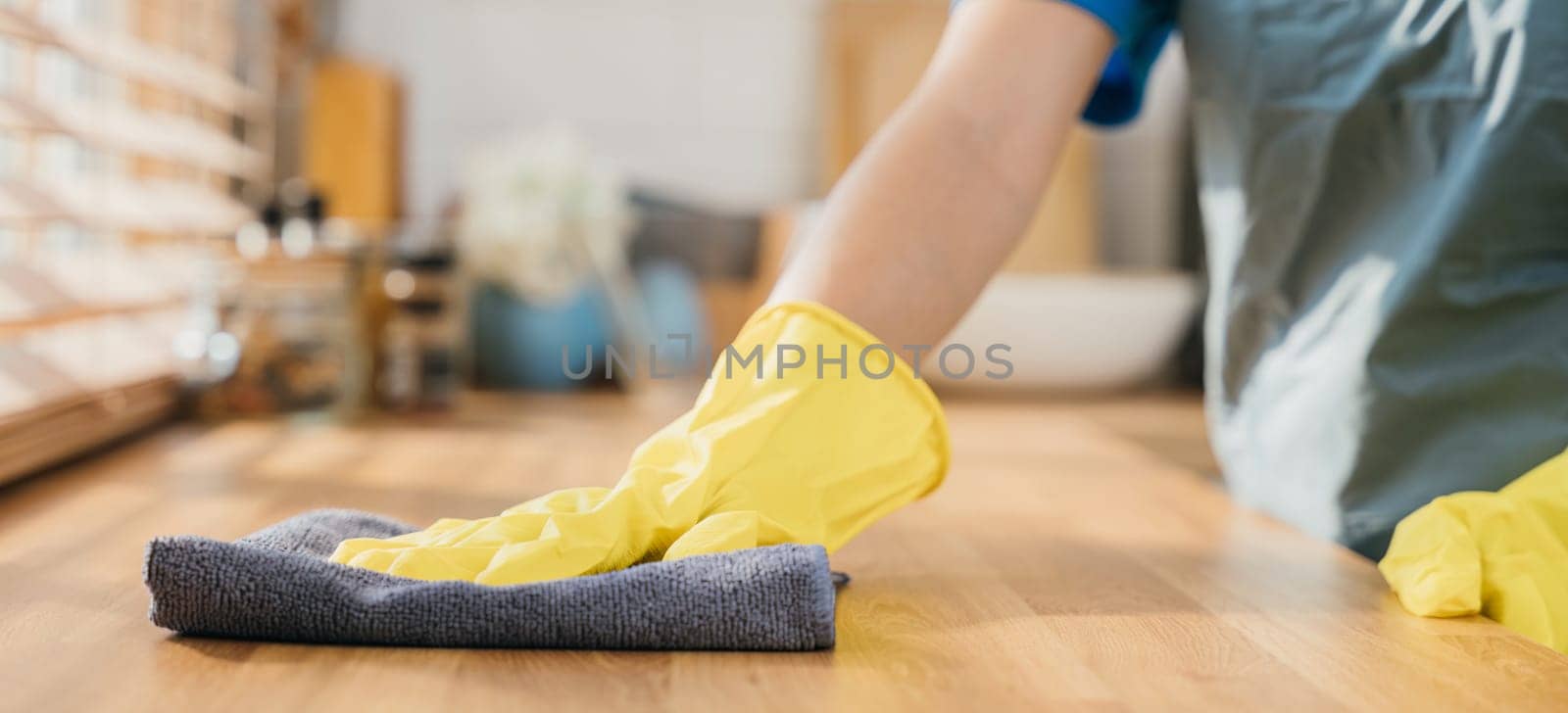 Housewife or maid in modern kitchen wipes dining table surface. Using professional cleaning products for home tidiness. Cleaner at work safety glove hygiene routine. Maid housekeeping concept. by Sorapop
