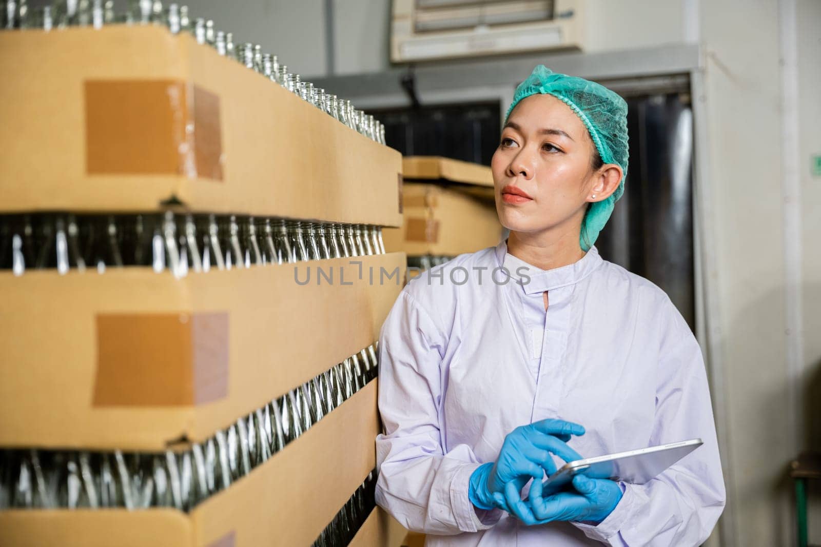 Within a beverage factory a QC woman checks products on the conveyor belt using a digital tablet. Her role involves quality control while examining the bottling line for liquid manufacturing.