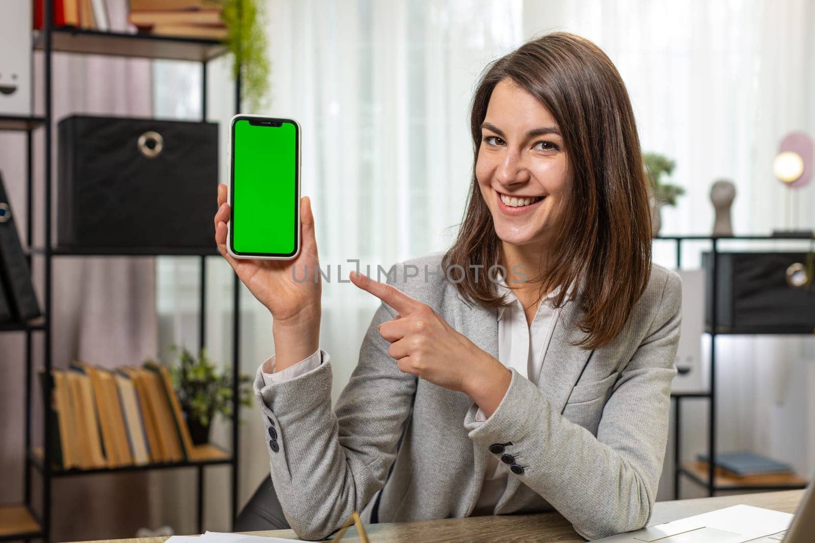 Happy smiling young businesswoman holding smartphone with green screen chroma key mock up recommend good application promotional sale offer. Freelancer woman girl at home office desk looking at camera