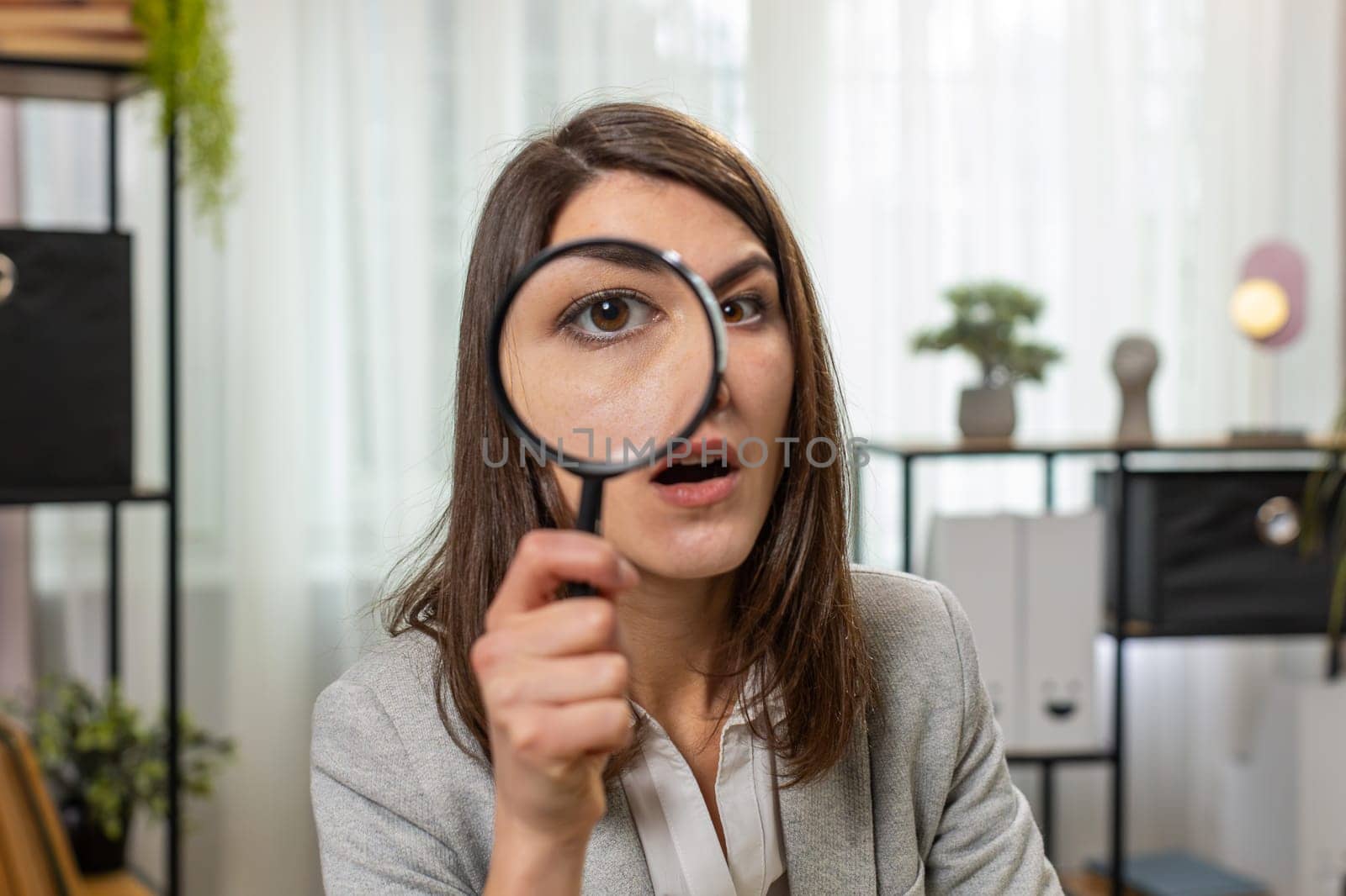 Investigator researcher businesswoman looking through magnifying glass with wow expression with big zoomed funny eye, searching, analyzing. Professional Caucasian freelancer girl looking at camera.