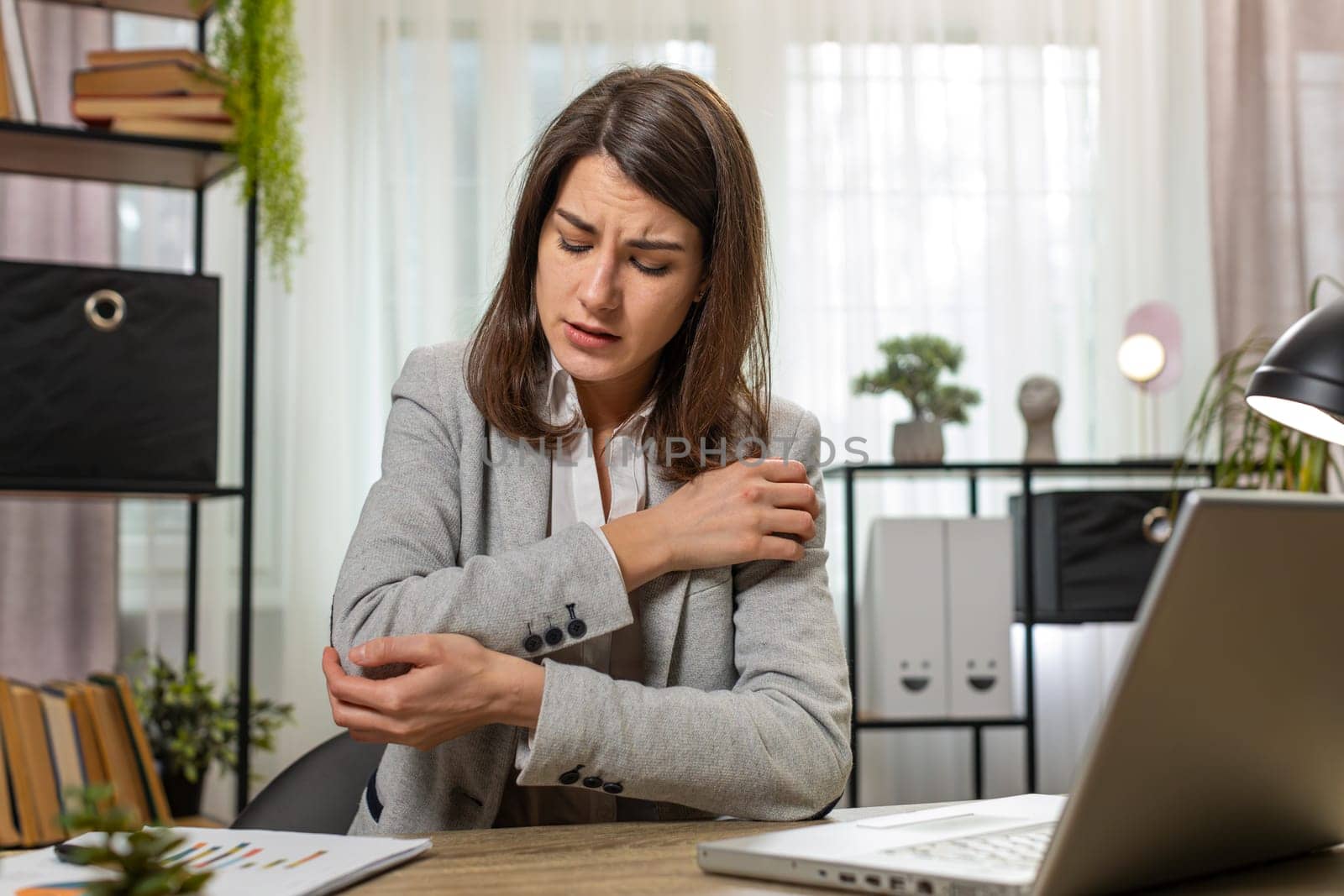 Tired Caucasian businesswoman girl sitting at home office desk with laptop and experiencing pain and discomfort in elbow joint and massaging painful hand. Arthritis, orthosis and bursitis concepts.