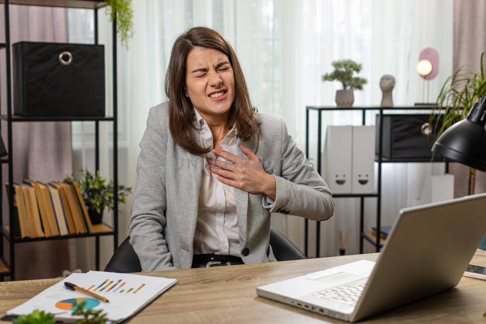 Stressed Caucasian young businesswoman using laptop and suffers from tension holding chest with heart pain attack risk of stroke symptom while working at home office desk. Girl freelancer at work.