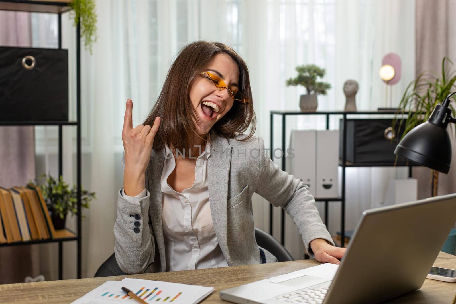 Happy excited Caucasian businesswoman working on laptop at home office desk putting on earbuds listening to favorite energetic disco music and dancing. Freelancer girl relaxing, taking a break.