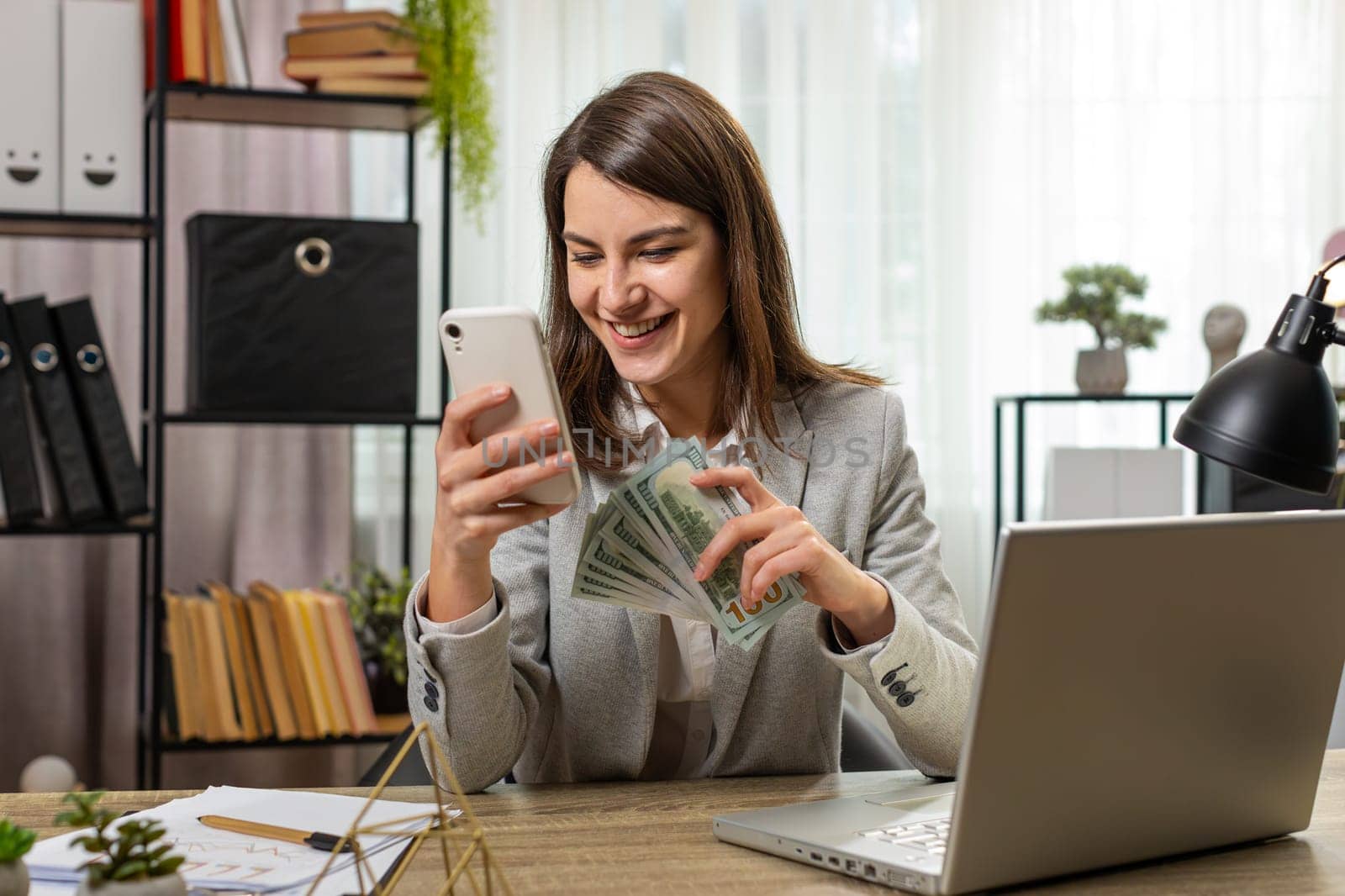 Happy rich business woman counting money cash, using smartphone calculator in home office. Successful Caucasian girl getting bundle of money in office calculates dividends cash money profits finances