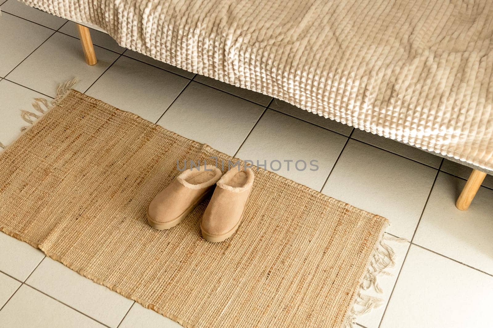 One pair of modern fashionable warm Uggs slippers lie in the center on a woven jute rug on a tiled floor near the sofa in the living room, close-up side view. Fashionable shoes concept.