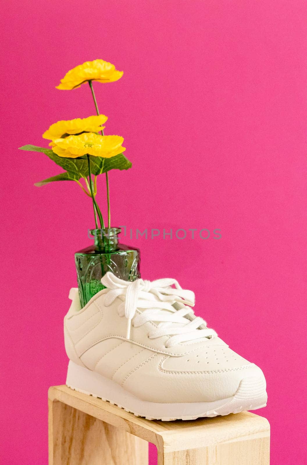 One modern fashionable white sneakers on a wooden box with a vase of yellow flowers in them stand on a lilac-pink background, close-up side view. Fashionable shoes concept.