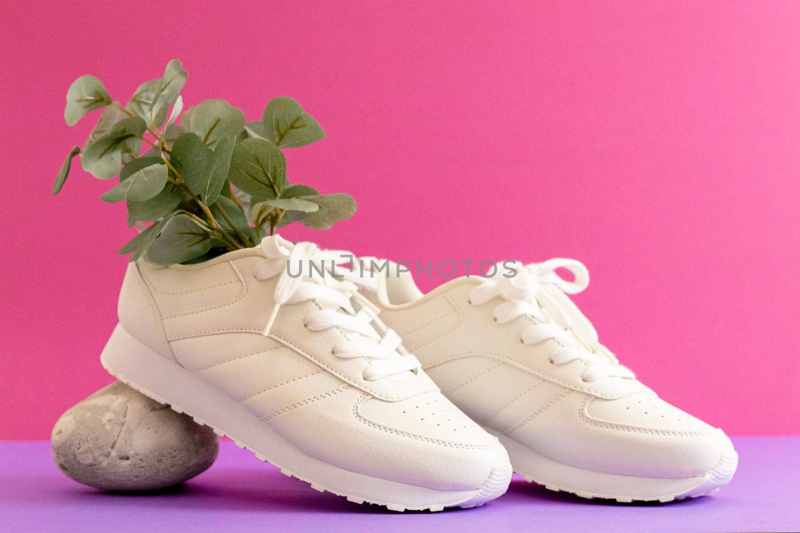 One pair of modern fashionable white sneakers from a eucalyptus branch rests on a gray stone on a lilac-pink background, close-up side view. Fashionable shoes concept.
