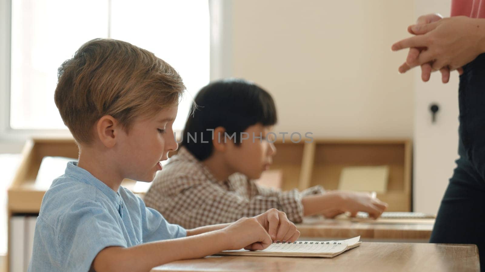Side view of caucasian elementary student looking a note and answer question while sitting in front of blackboard. Young teacher explain classwork or test while children attend the class. Pedagogy.