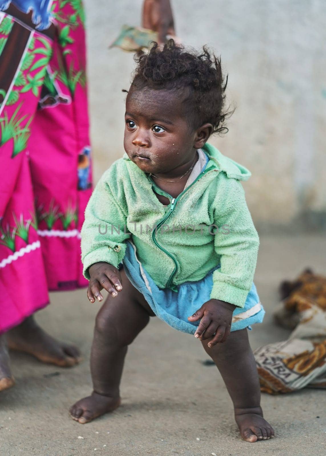 Ranohira, Madagascar - April 29, 2019: Unknown little Malagasy kid, barefoot, barely standing, mother stands near. People in Madagascar are poor but cheerful especially children