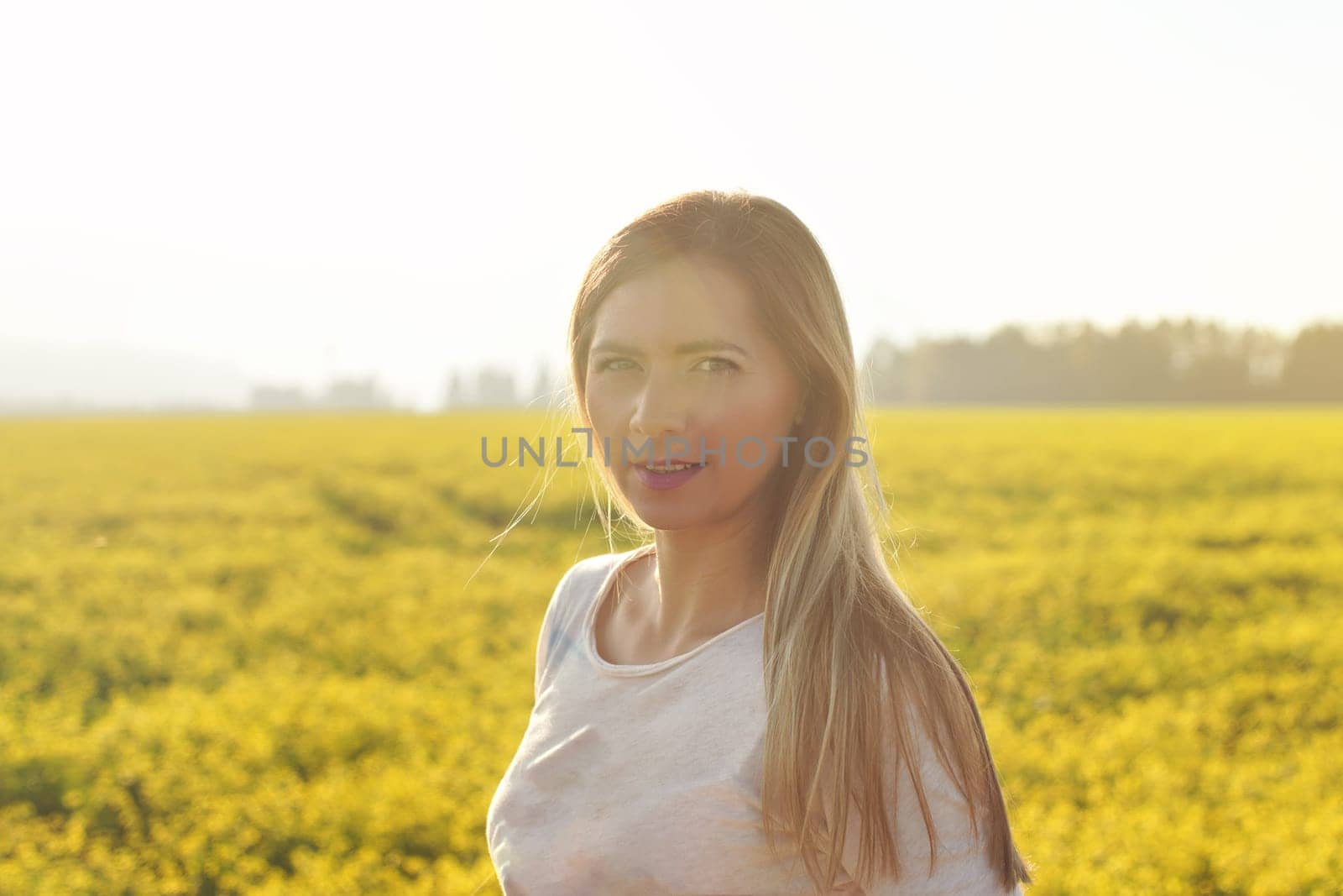 Portrait of young woman with long hair in strong afternoon backlight sun, blurred field with yellow flowers background by Ivanko