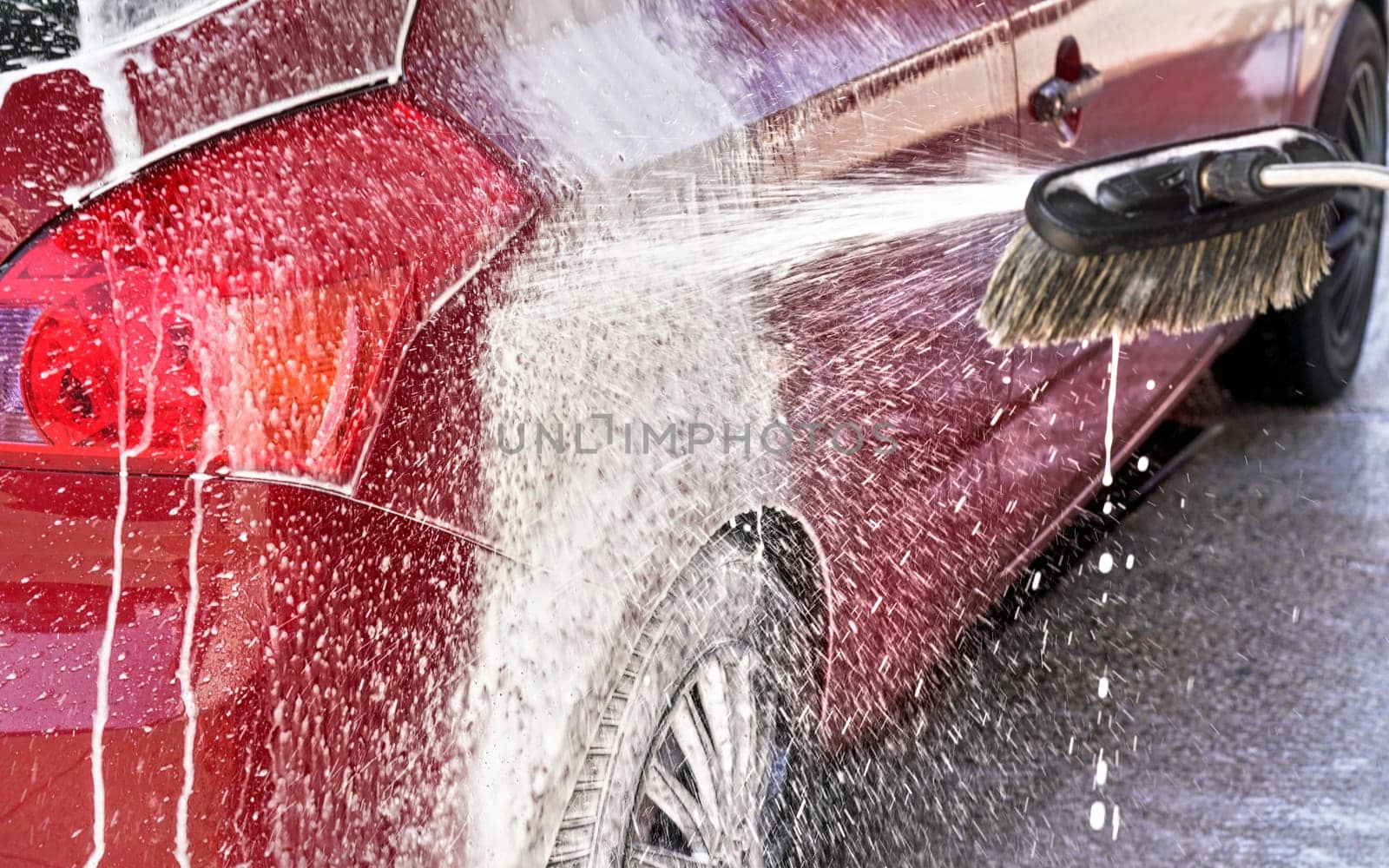 Red car washed in self serve carwash, detail on white soap foam spraying from brush / broom onto side above rear wheel by Ivanko