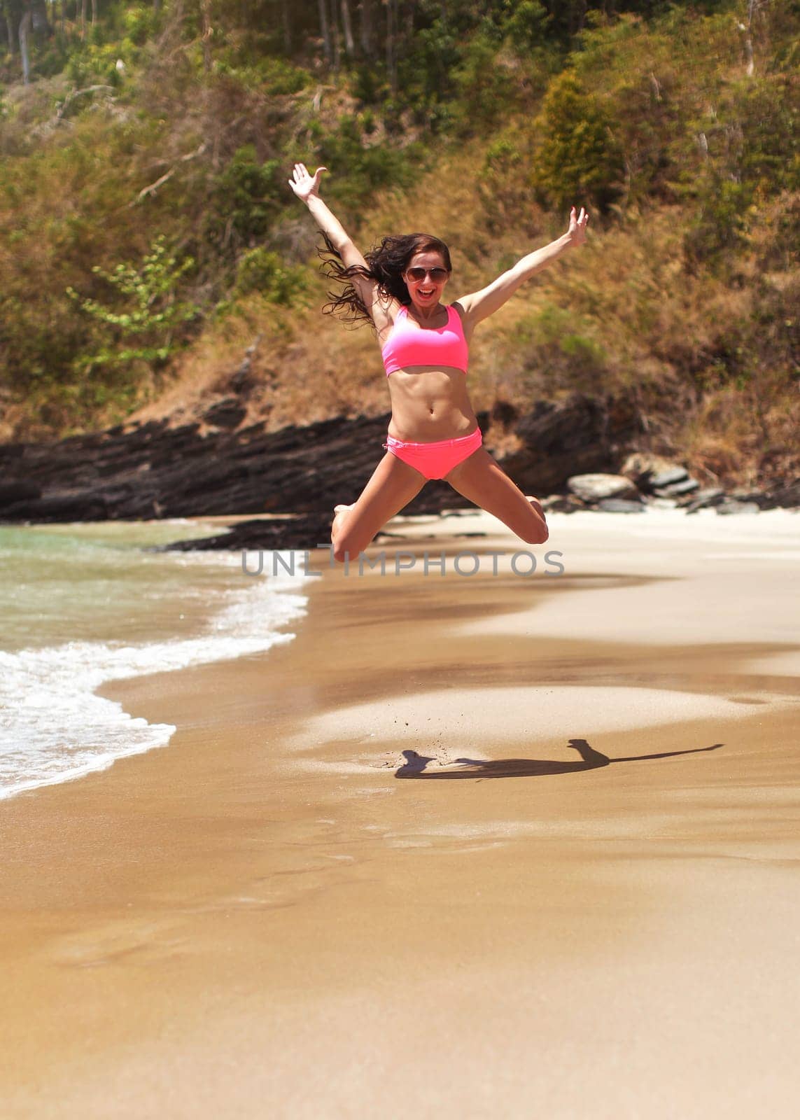 Young woman in pink bikini, wearing sunglasses, jumping high over smooth sand beach, her arms spread - looking happy