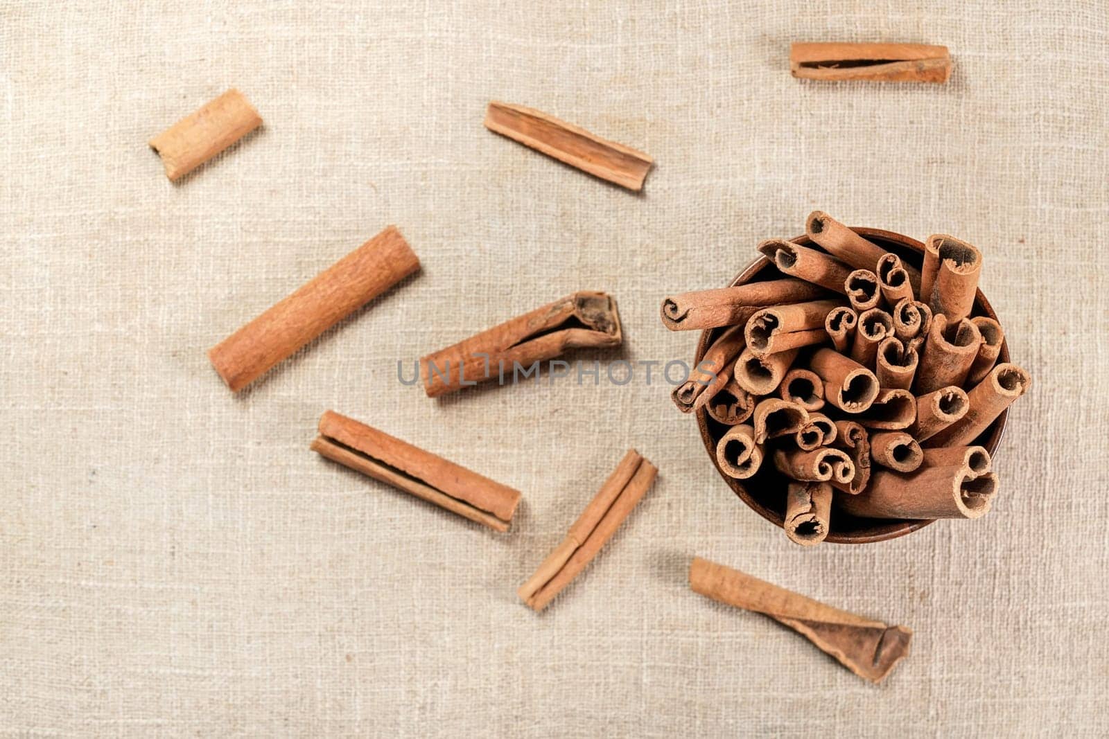 Closeup detail - Cinnamon bark sticks in small wooden cup, some scattered on linen tablecloth view from above by Ivanko