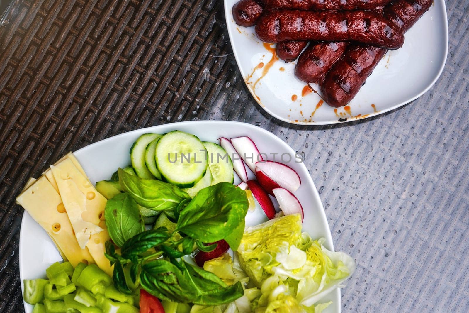 Two white plates, one with grilled bratwursts / frankfurters, other with salad - cucumbers, radish, basil, pepper and cheese. View from above by Ivanko