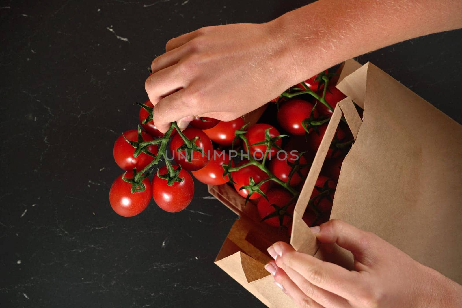 Taking out red cherry tomatoes with green leaves from brown paper bag over dark marble like board, detail on woman hand holding fruits by Ivanko