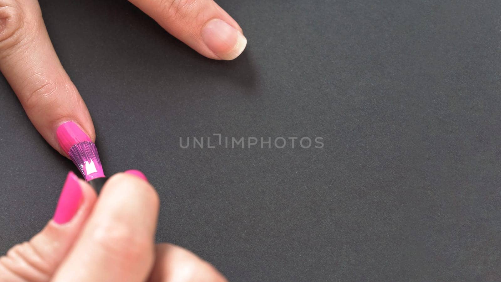 Applying pink nail polish - closeup photo of woman hands, little brush and fingernails on gray / black background, space for text right side by Ivanko