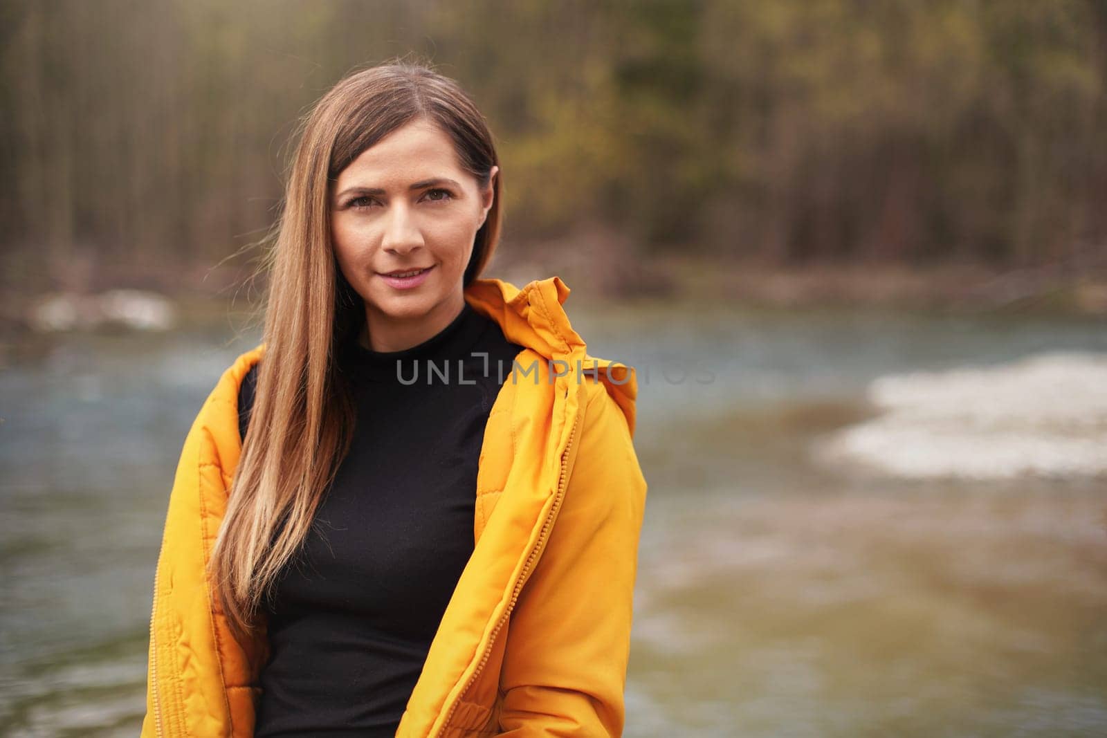 Portrait of young woman in yellow jacket, smiling, long hair down, blurred trees and river background