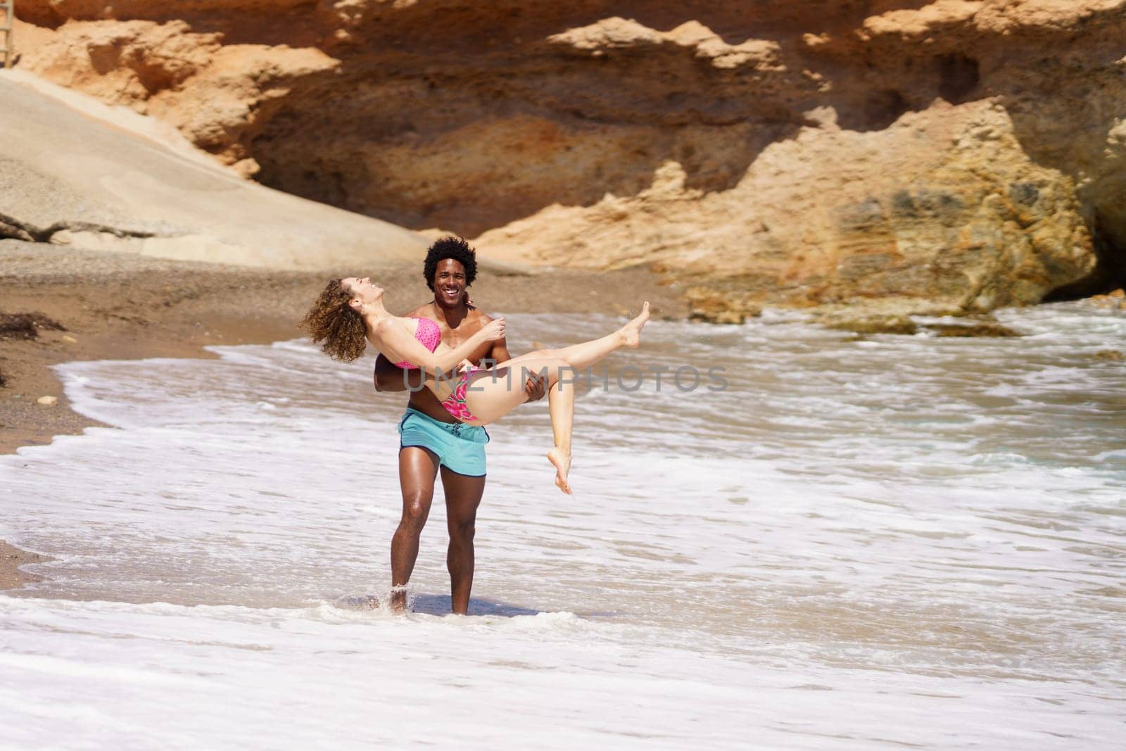 Full body of cheerful African American man smiling and raising woman in swimwear while walking on wet beach during holidays