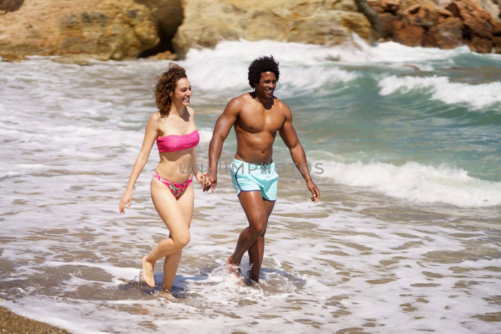 Full body of cheerful multiracial couple holding hands and strolling together on coast while enjoying summer trip in sea