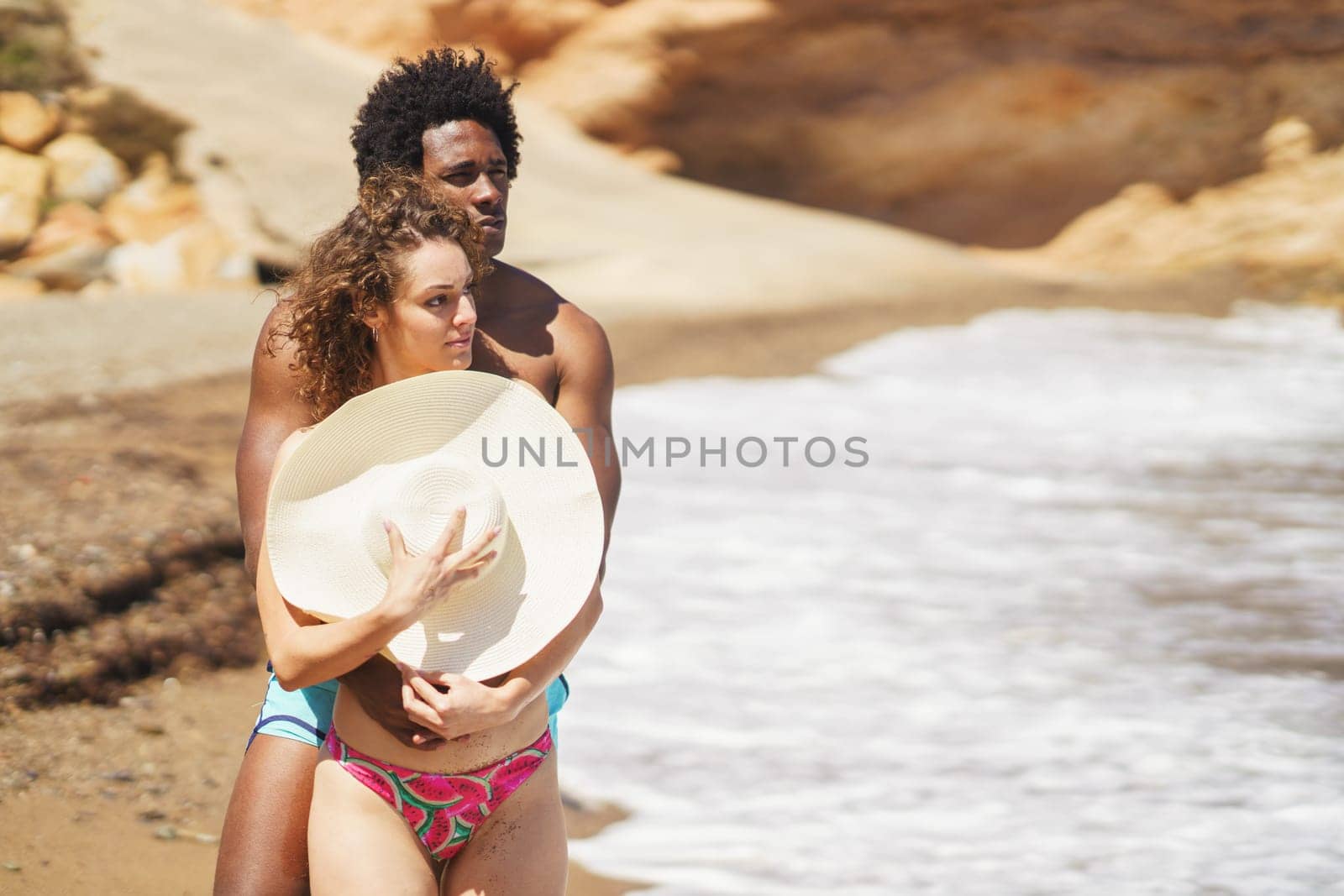 Young African American man with Afro hairstyle hugging girlfriend from behind standing on sandy beach, covering breast with hat while spending time together near waving sea