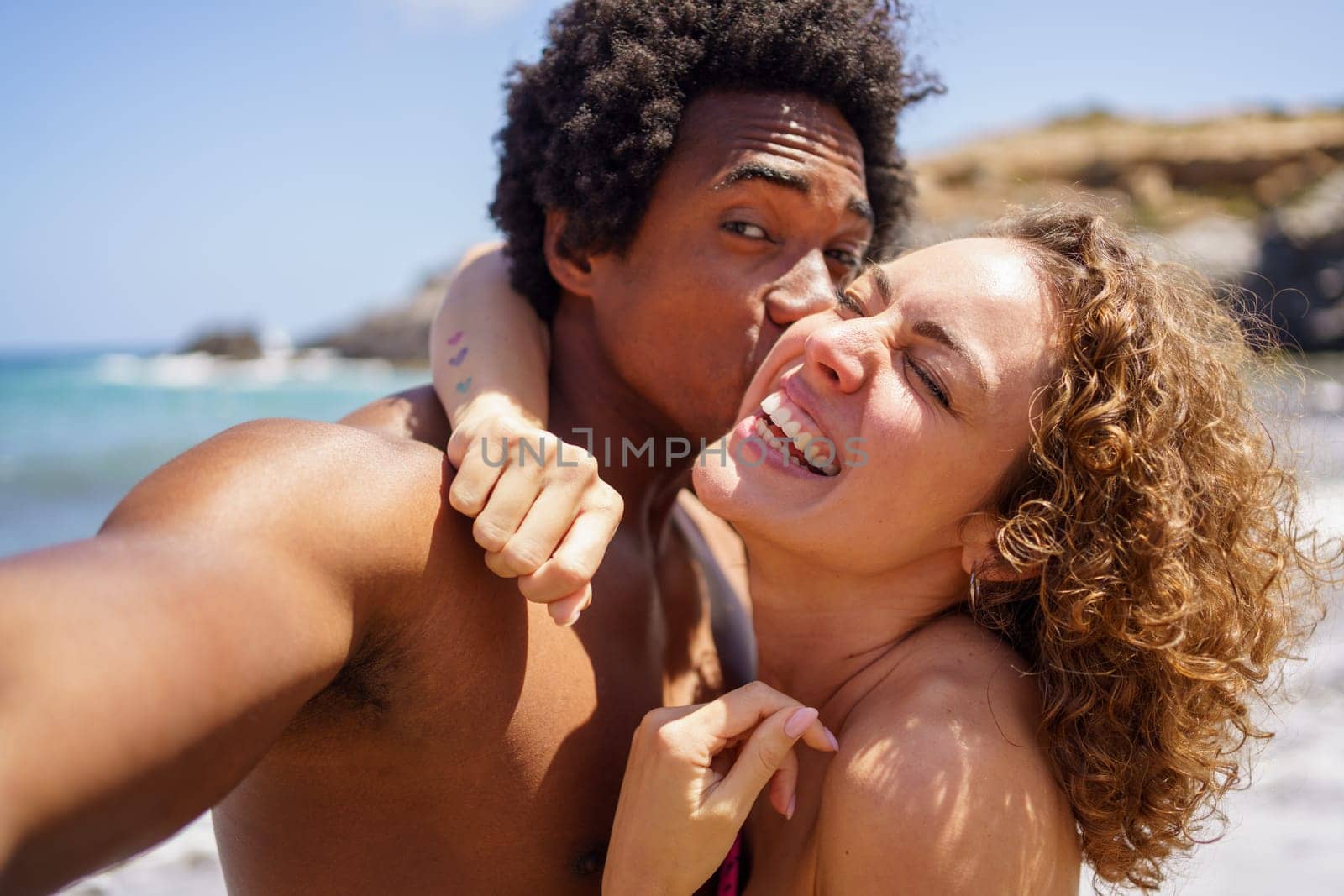 Loving African American man with Afro hair taking selfie while kissing girlfriend on cheek during summer vacation near sea