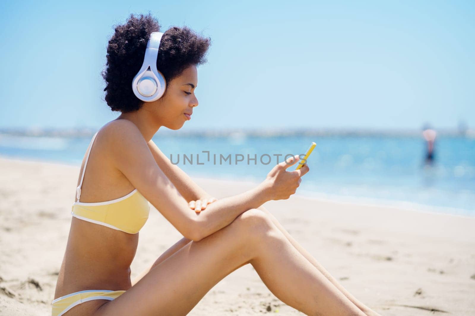 Side view of content young fit African American female millennial with curly hair, in trendy swimwear browsing smartphone, and listening to music in headphones sitting on sandy beach near sea