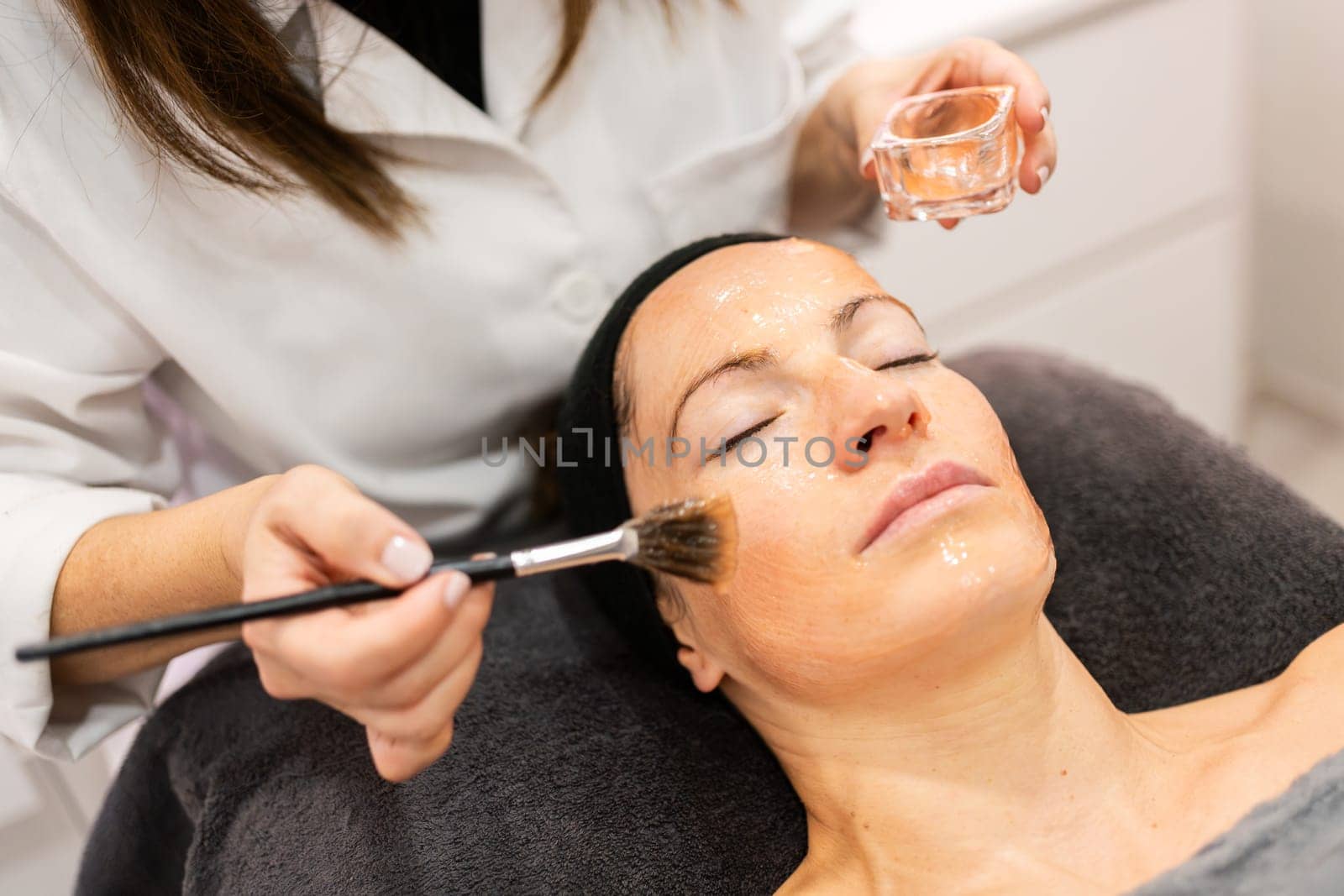 Unrecognizable cosmetologist in white uniform using brush and applying cosmetic product on face of female customer during facial beauty procedure in salon