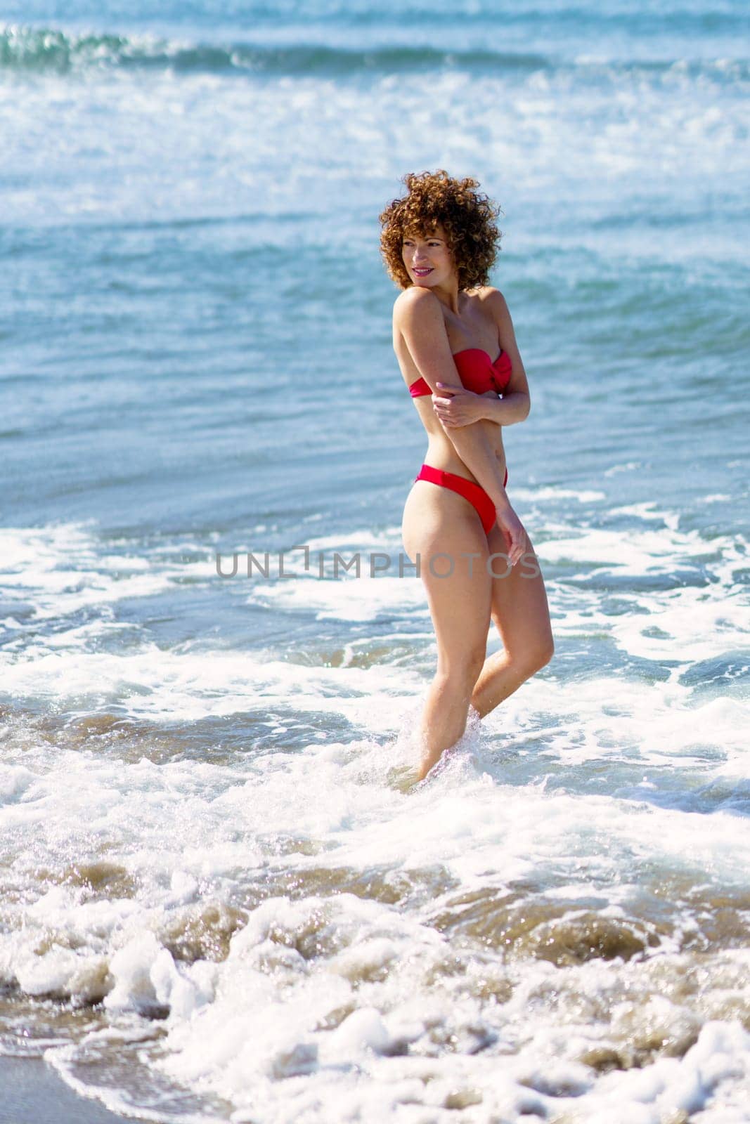 Side view of confident graceful young female with curly hair in red bikini smiling and looking over shoulder, while standing in sea water near sandy beach on sunny day