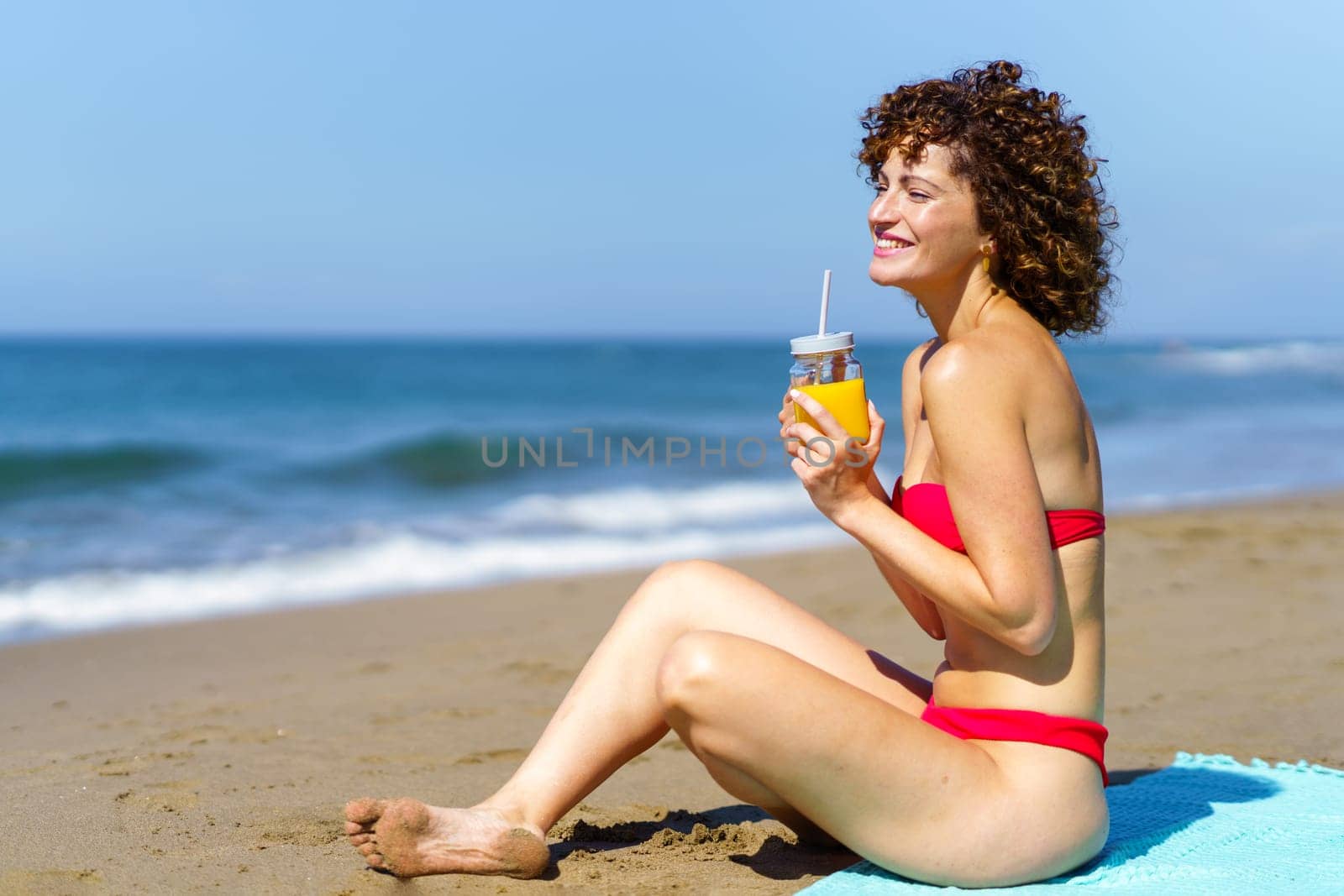 Side view of young female with curly hair and in bikini sitting on sandy beach with glass of orange juice and looking away