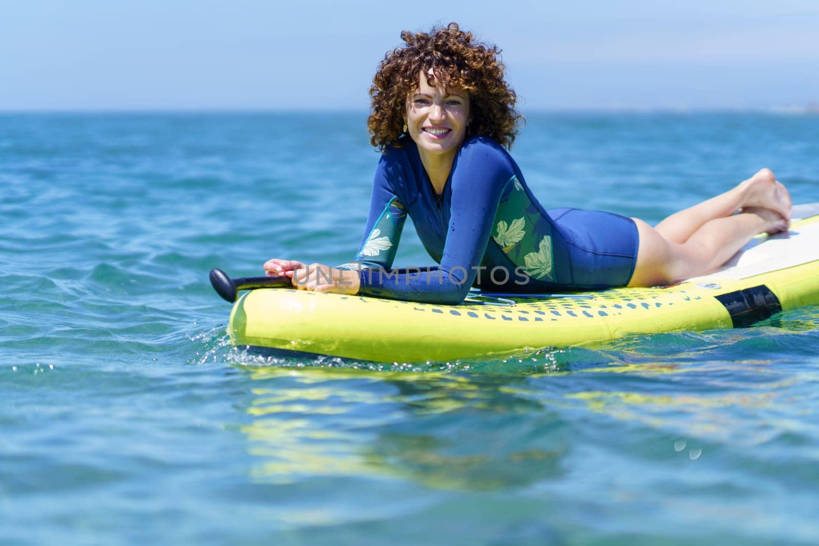 Smiling curly lady in wetsuit resting on paddleboard on surface of sea and looking at camera while enjoying daylight