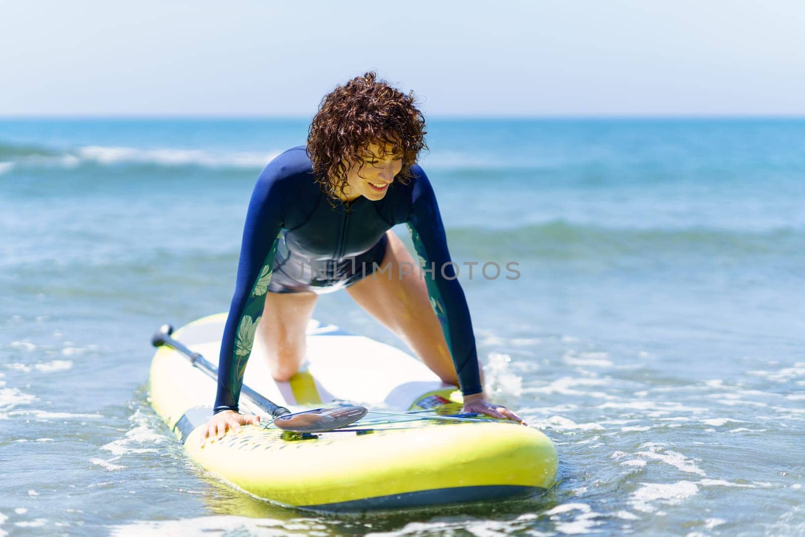 Cheerful young woman in wetsuit with curly hair enjoying surfing on SUP board while floating on sea shore during summer vacation
