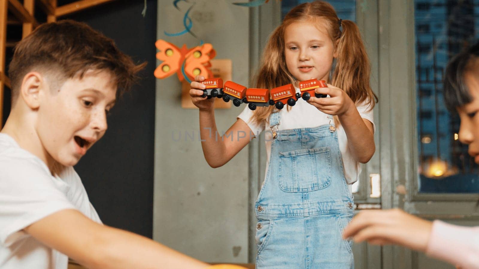 Girl playing wooden train while diverse student sitting at play room. Boy looking at lovely cute student hold toy while his friend talking about lesson in break time. Creative activity. Erudition.