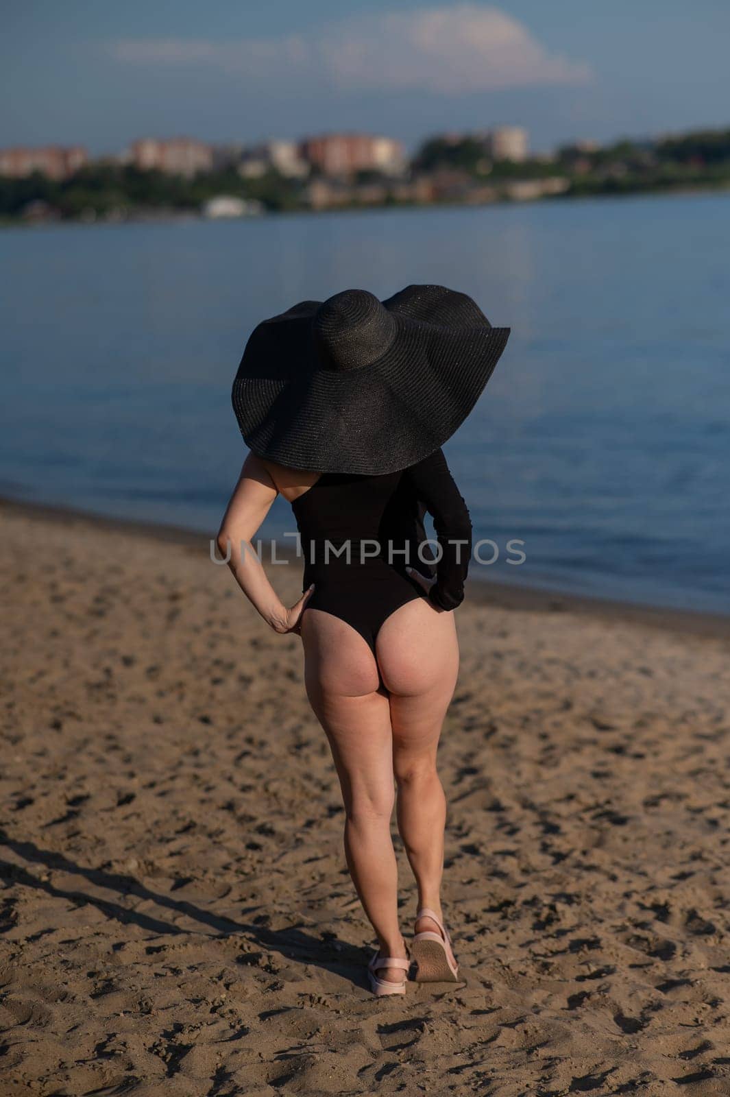 Rear view of woman in large straw hat and black swimsuit posing on the beach. by mrwed54