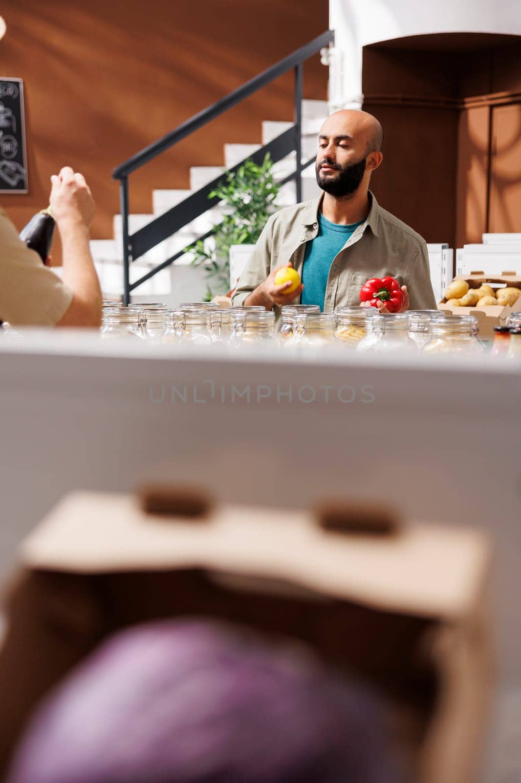 Middle Eastern male customer holds a lemon in one hand and red pepper in another, admiring them both. Young bearded client browsing in an environmentally friendly grocery store.