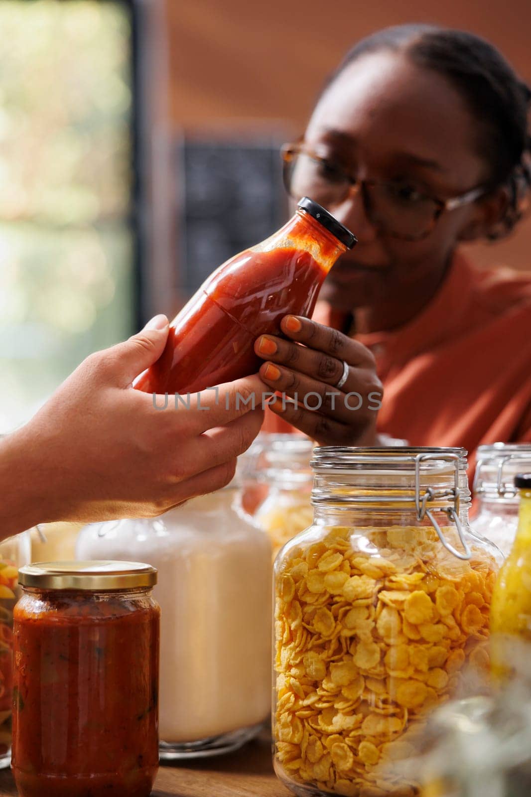 Customer shopping for vegan sustainable food items in a zero waste shop with vendor providing information. Environmentally friendly woman analyzing homemade sauces ideal for healthy eating.