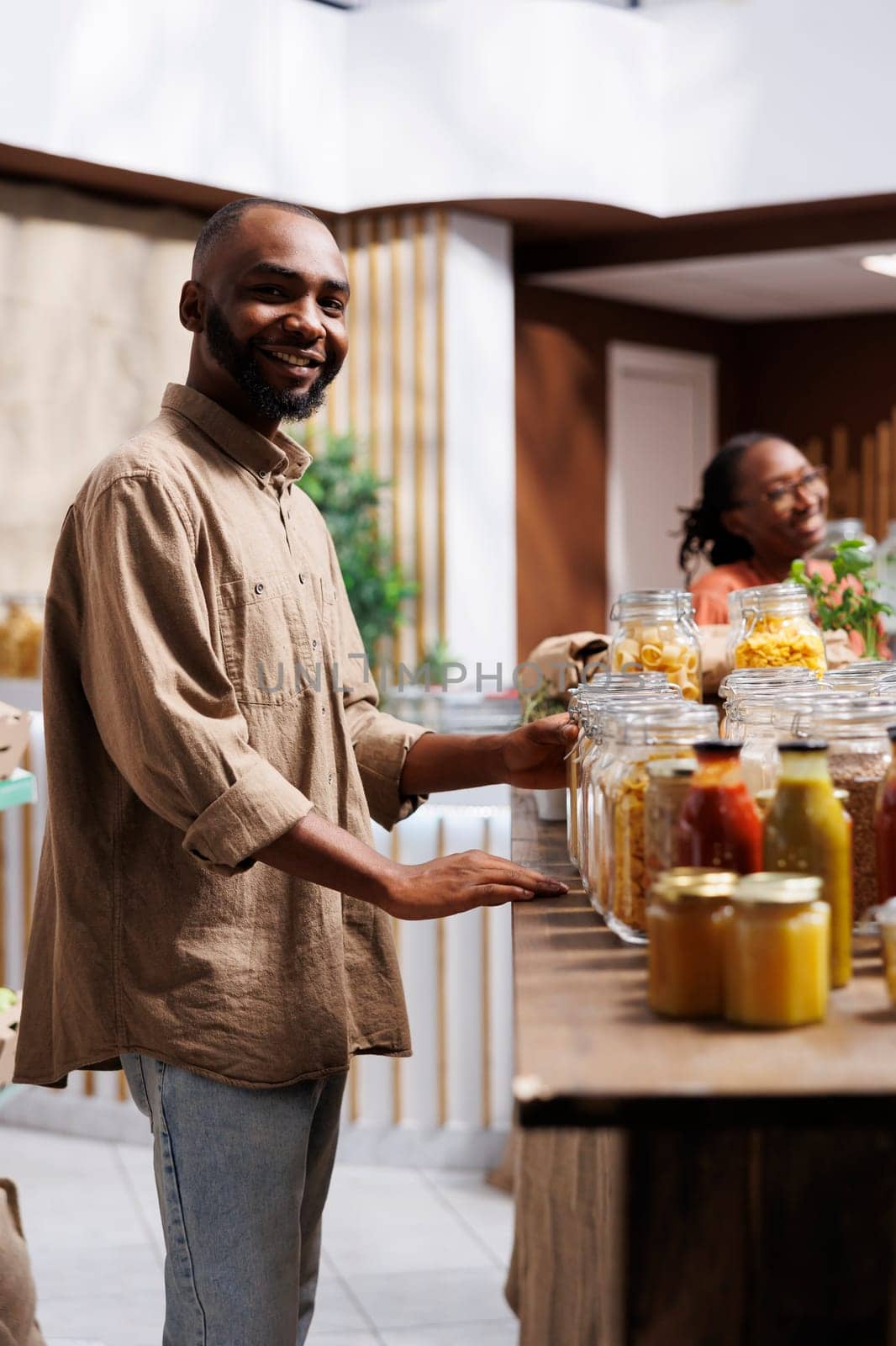 In a modern grocery shop, a happy African American man looks at the camera. The shelves feature reusable packaging, eco friendly products, and a selection of items.