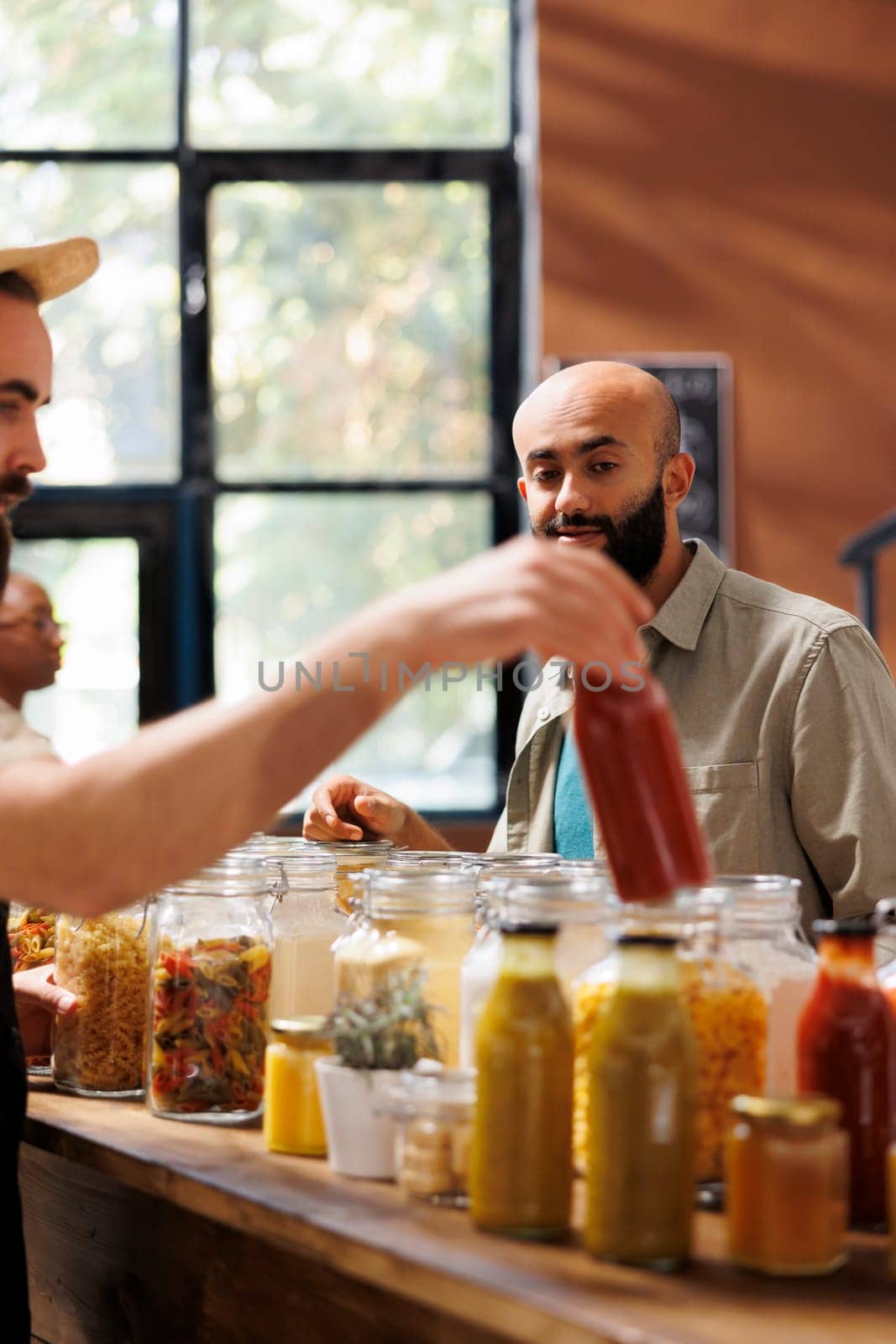 Middle Eastern customer looks at glass bottle filled with organic sauce being held by storekeeper. Local vendor providing male client with bulk natural sustainable products during an event.