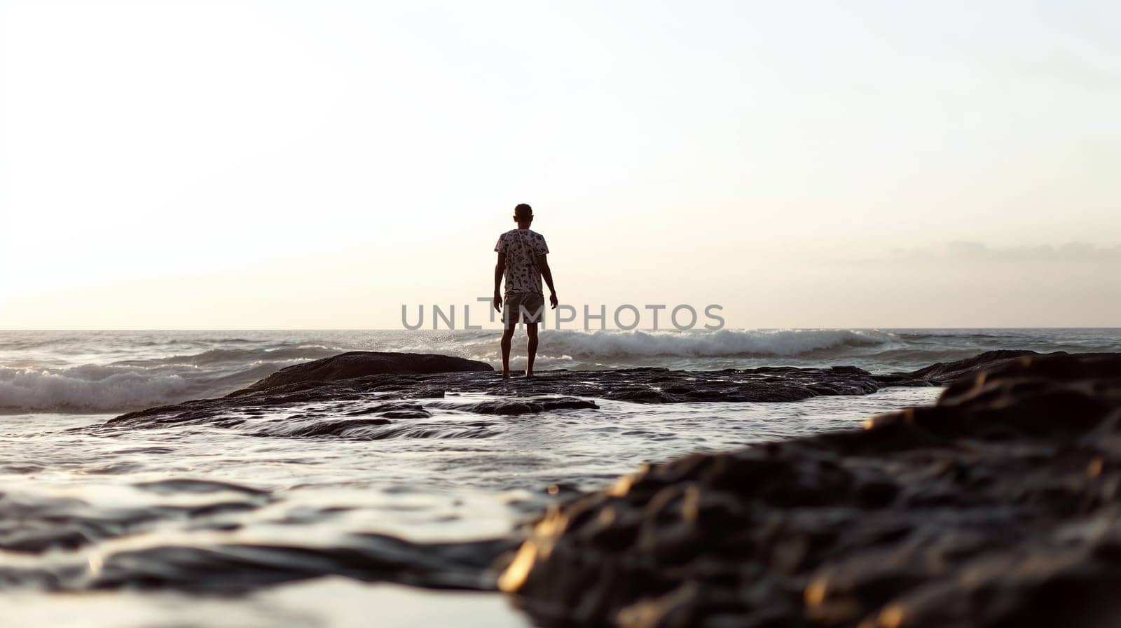 A lone man is captured walking on the wet rocks beside the ocean waves, with the sunset casting a soft glow in the background - Generative AI