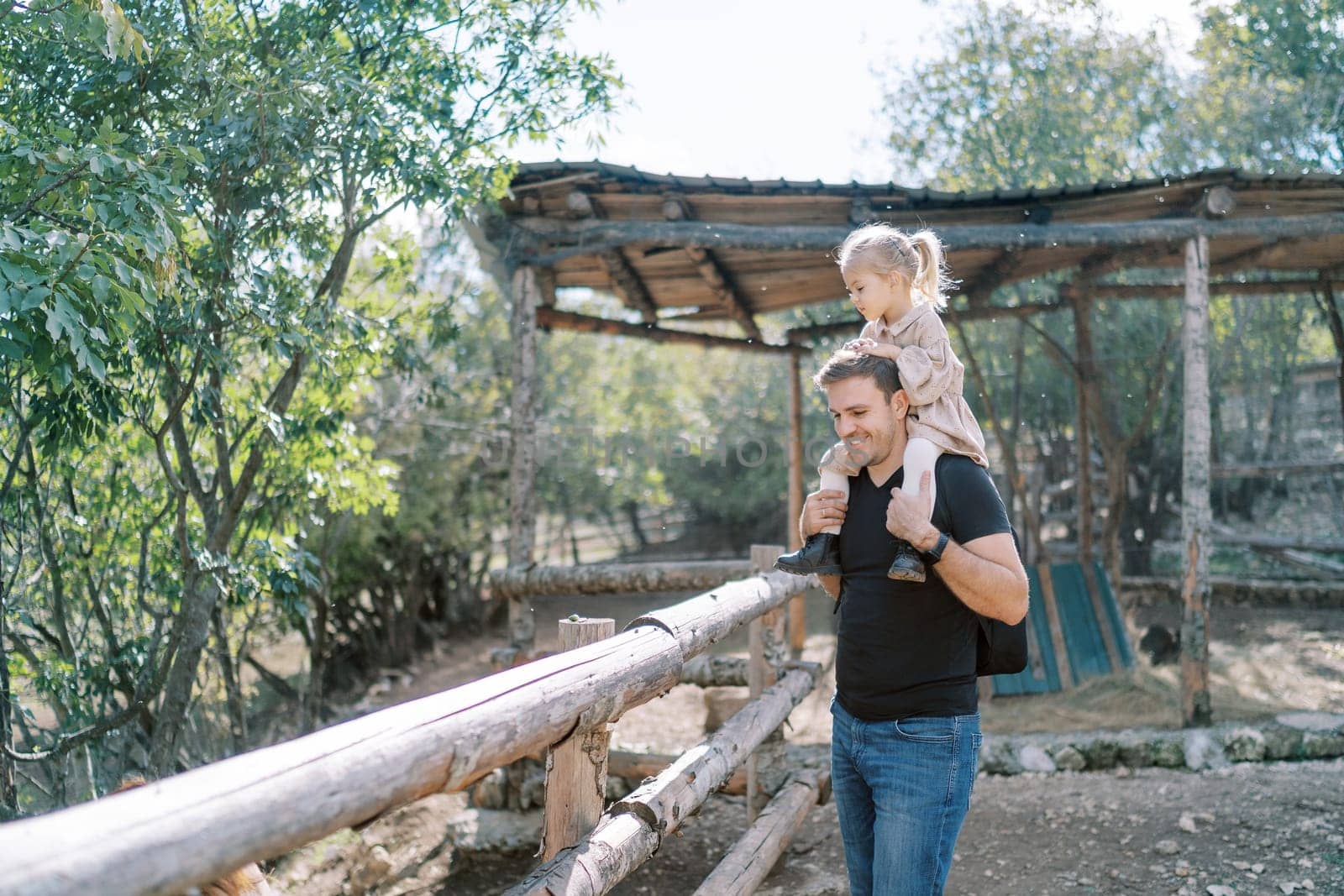 Smiling dad with little girl on shoulders standing near wooden fence of corral in park. High quality photo