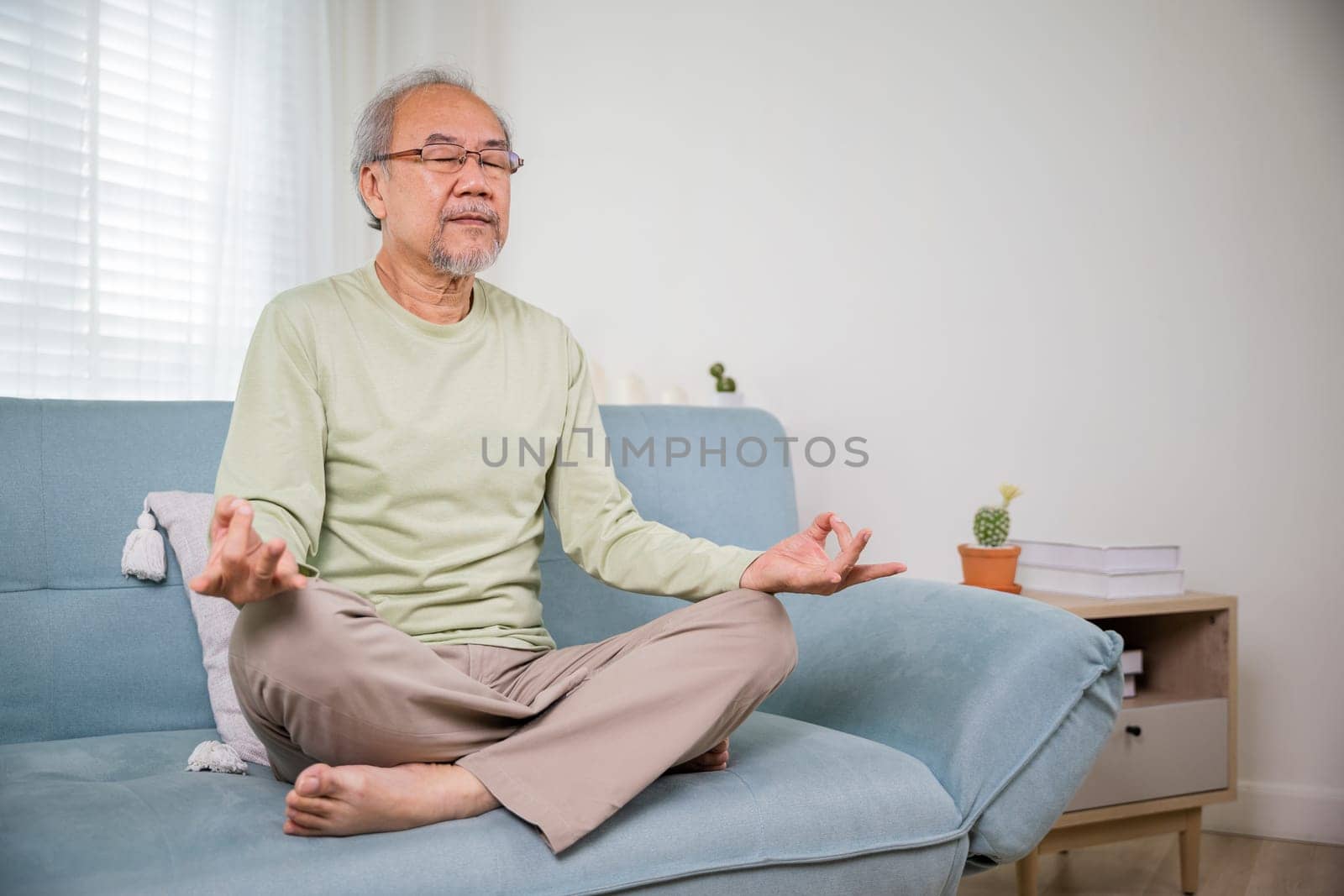 Lifestyle senior man sitting on sofa in living room holding hands in mudra practicing home yoga in lotus pose, Elderly relaxed man do physical exercises following healthy on stress free weekend