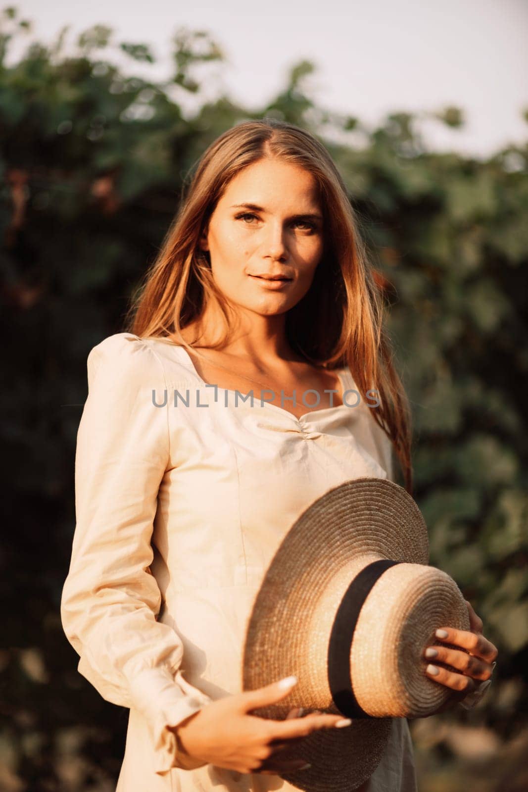 Woman with straw hat stands in front of vineyard. She is wearing a light dress and posing for a photo. Travel concept to different countries by Matiunina