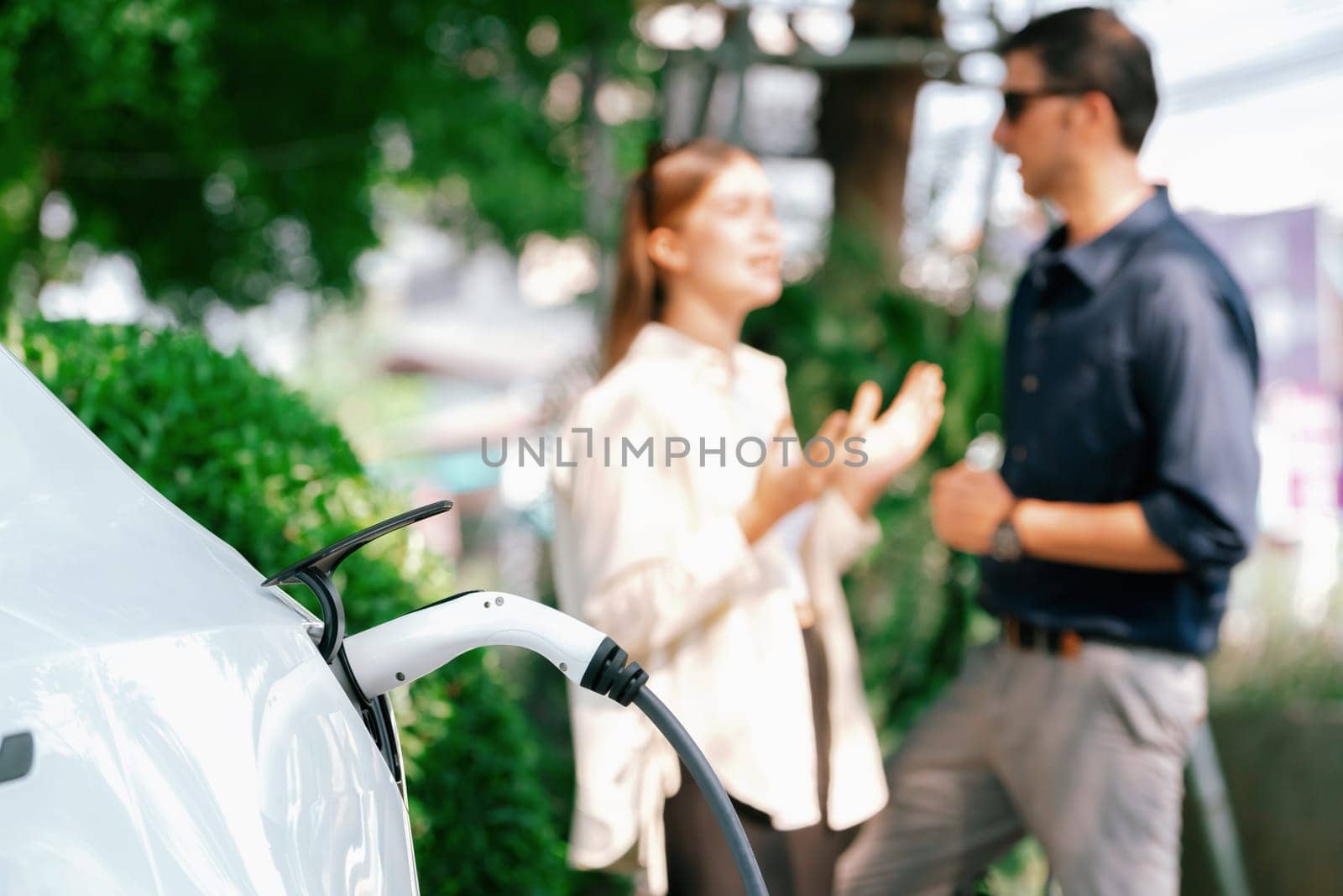 Young couple recharge electric car's battery from charging station in outdoor green city park in springtime. Rechargeable EV car for sustainable environmental friendly urban travel lifestyle.Expedient