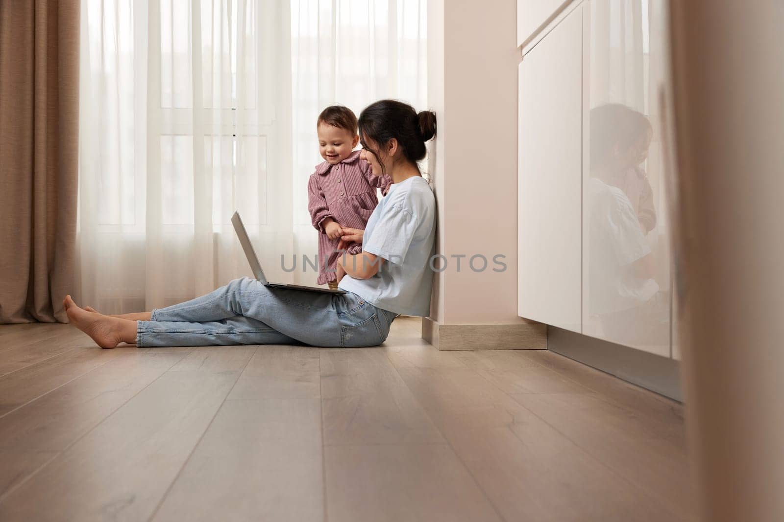 woman sitting on the floor and working on laptop with her little child girl at home by erstudio