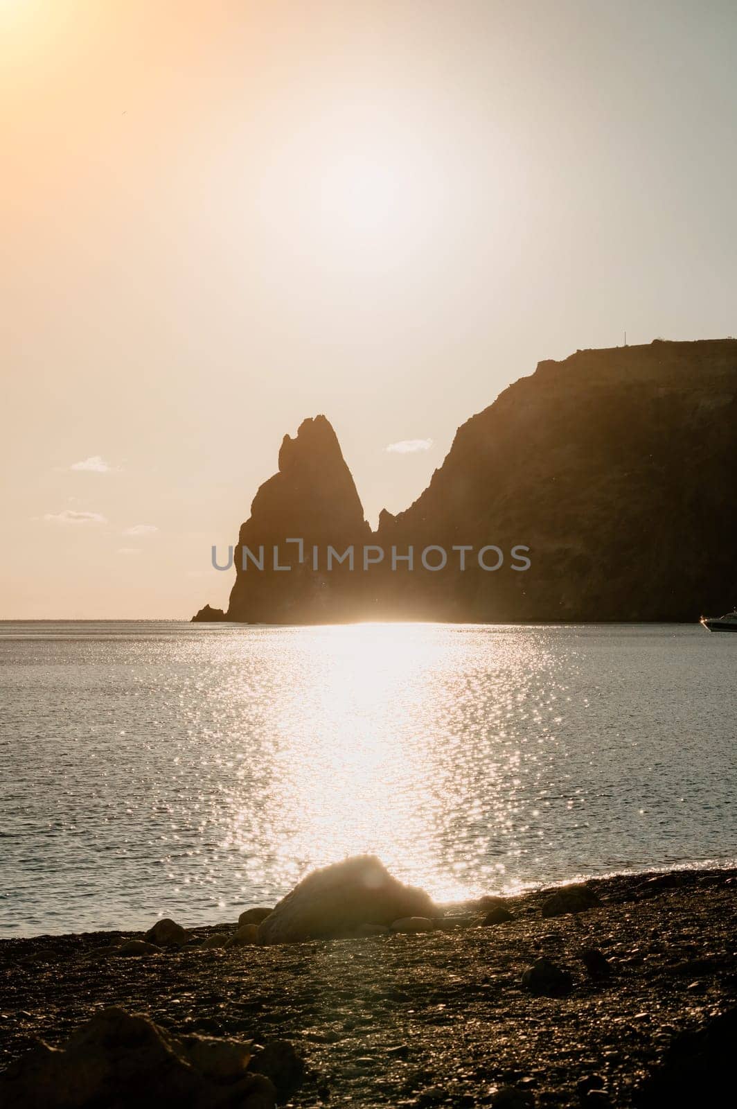 A red burning sunset with the silhouette of a cliff over the sea. Abstract nature summer or spring ocean sea background. Small waves on golden warm water surface with bokeh lights from sun.